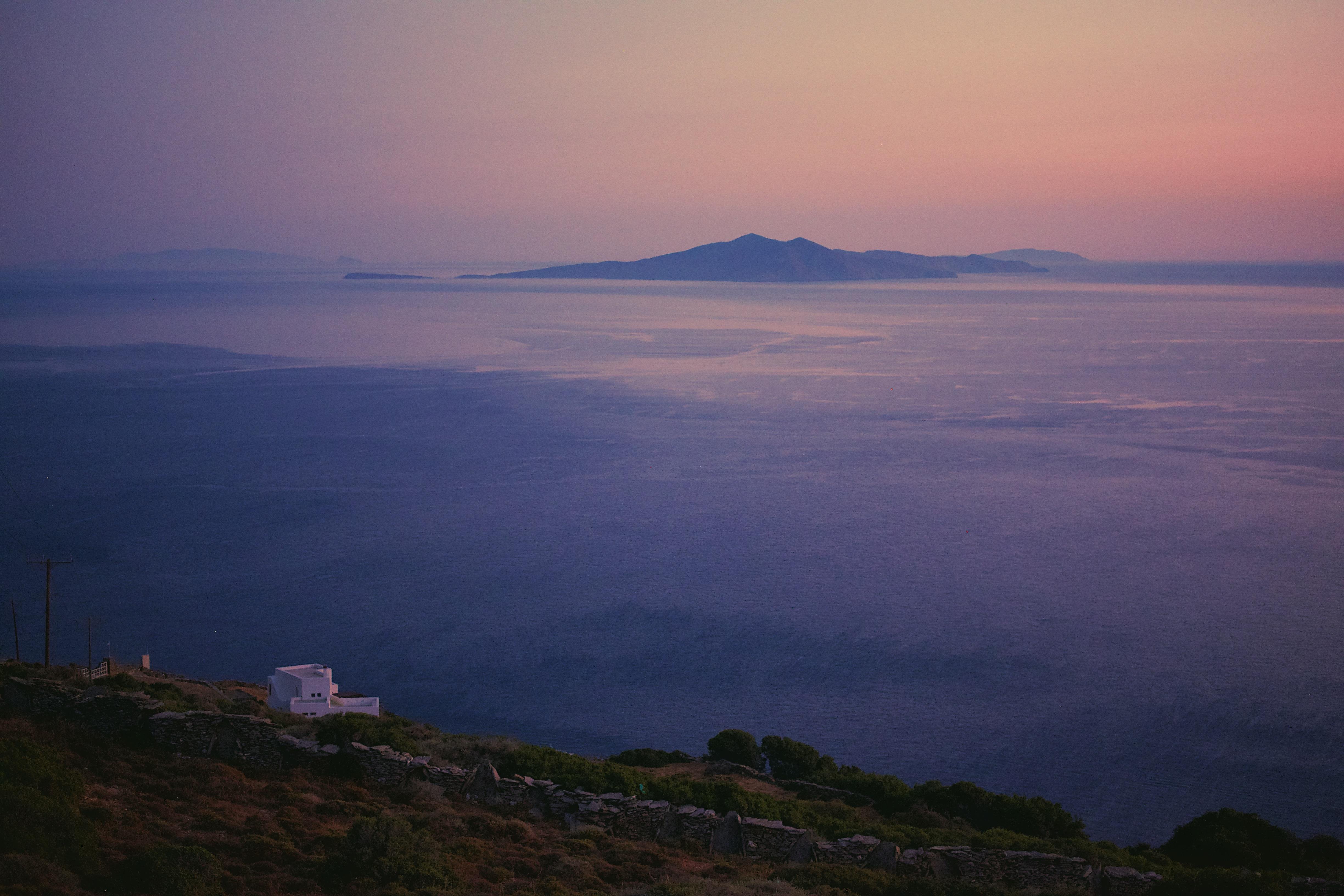 Coastal landscape at sunset with a distant island on the horizon. The sea appears calm, reflecting hues of purple and pink from the sky. A white building is situated on a hill in the foreground, surrounded by sparse vegetation.