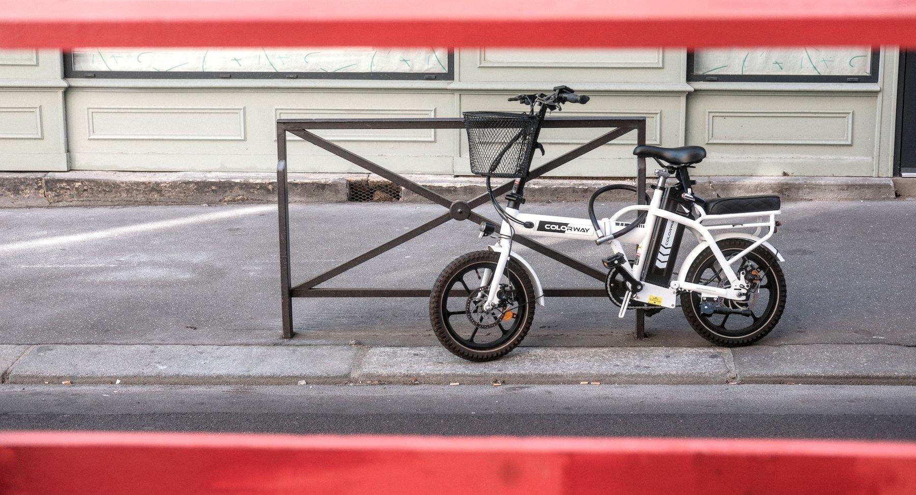 Photo horizontale. Vue entre 2 barreaux rouges horizontaux sur un petit vélo blanc attaché à une barrière urbaine marron sur un trottoir