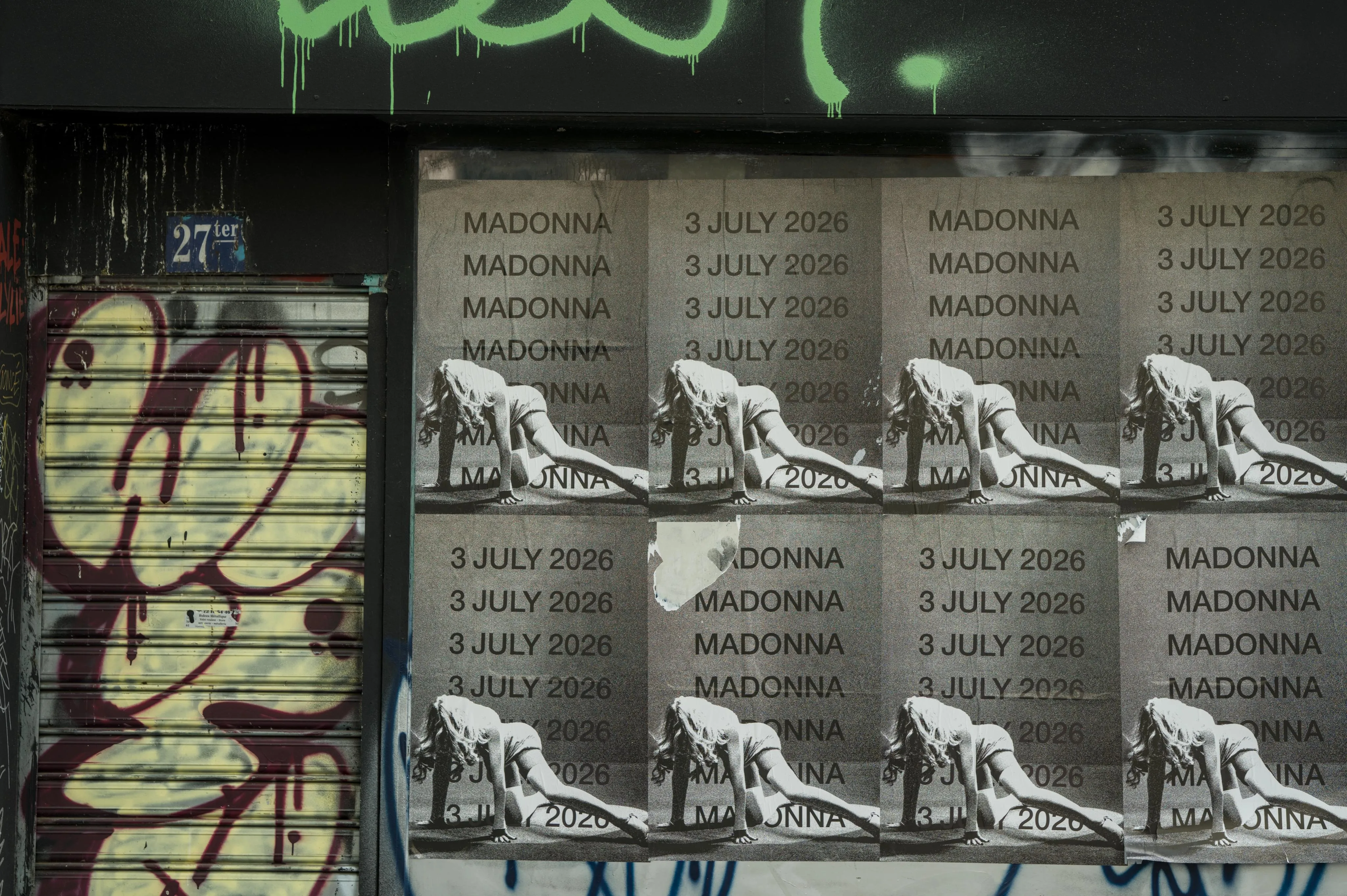 Photograph of a closed-down store front with an iron curtain on the door painted with graffiti and Madonna posters. 
There are 2 rows of 4 Madonna posters, all with the same photograph of Madonna dancing on the ground. Every second Madonna poster has the words MADONNA MADONNA MADONNA repeated in a column, every other one has the date repeated in a column: 3 JULY 2026.
One of the Madonna posters has a torn down corner.