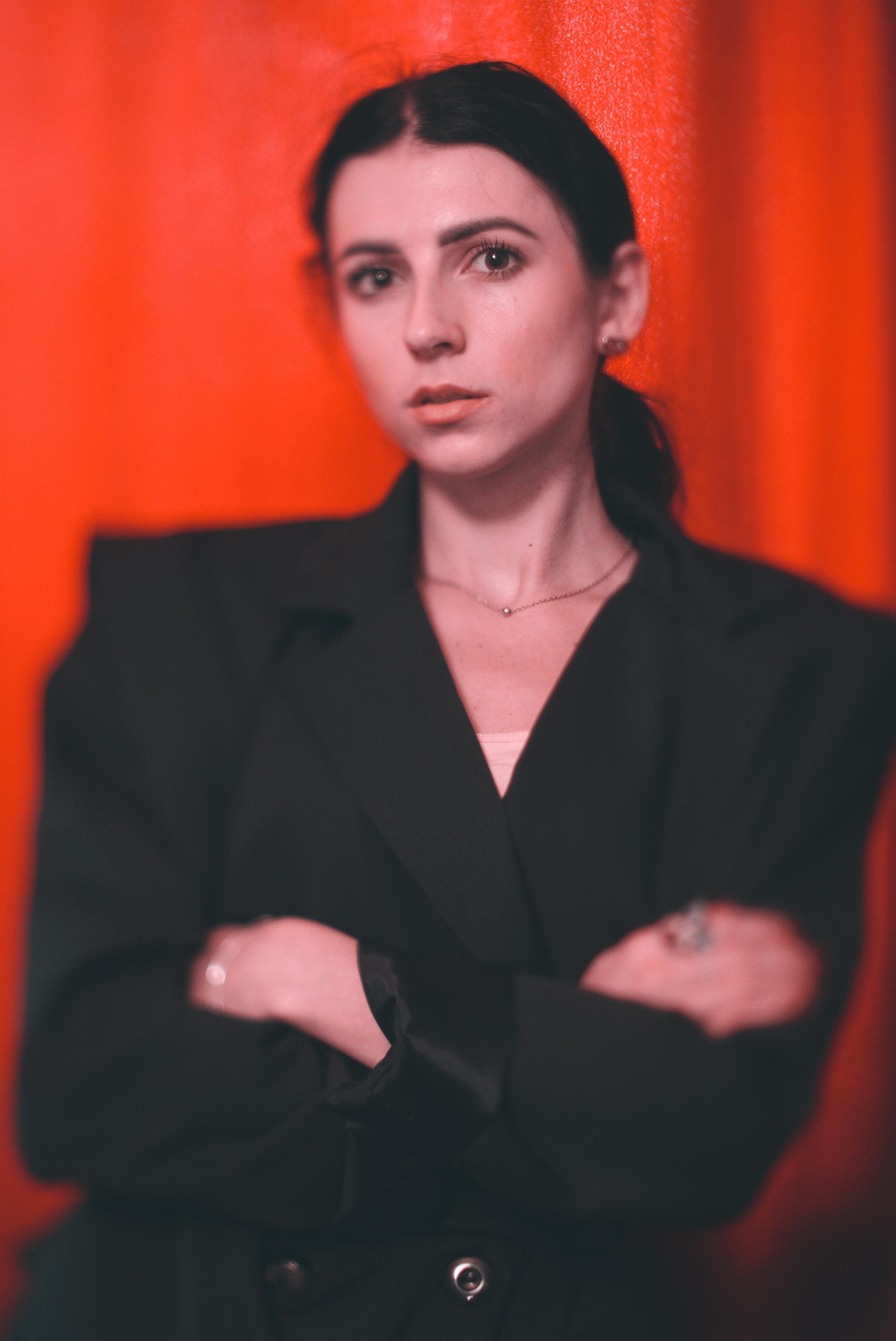 Woman with dark hair and a serious expression stands with crossed arms in front of an orange-red curtain. She wears a black blazer, a light necklace, and has small earrings.