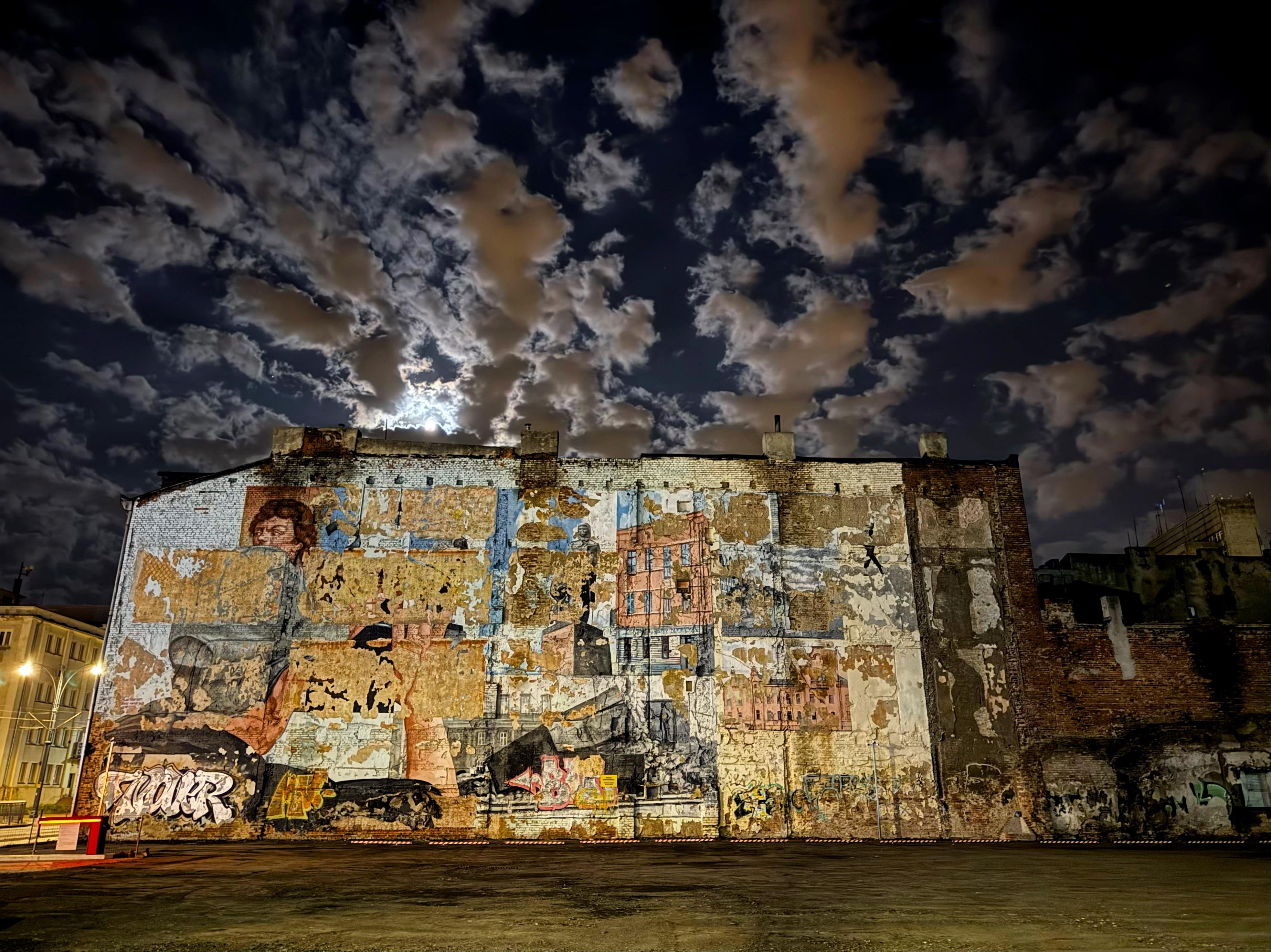 The image showcases a large wall covered with a complex mural. The mural itself appears to be weathered and partially eroded, with various images and colors combined in a collage-like manner. There are signs of graffiti and patches of different painted artwork, some of which are faded or peeling away from the surface. The wall is part of a larger building, and the scene is set at night under a sky with dramatic cloud formations illuminated by moonlight. The atmosphere of the image is somewhat surreal and moody due to the nighttime setting and the eclectic mix of art and decay on the wall.