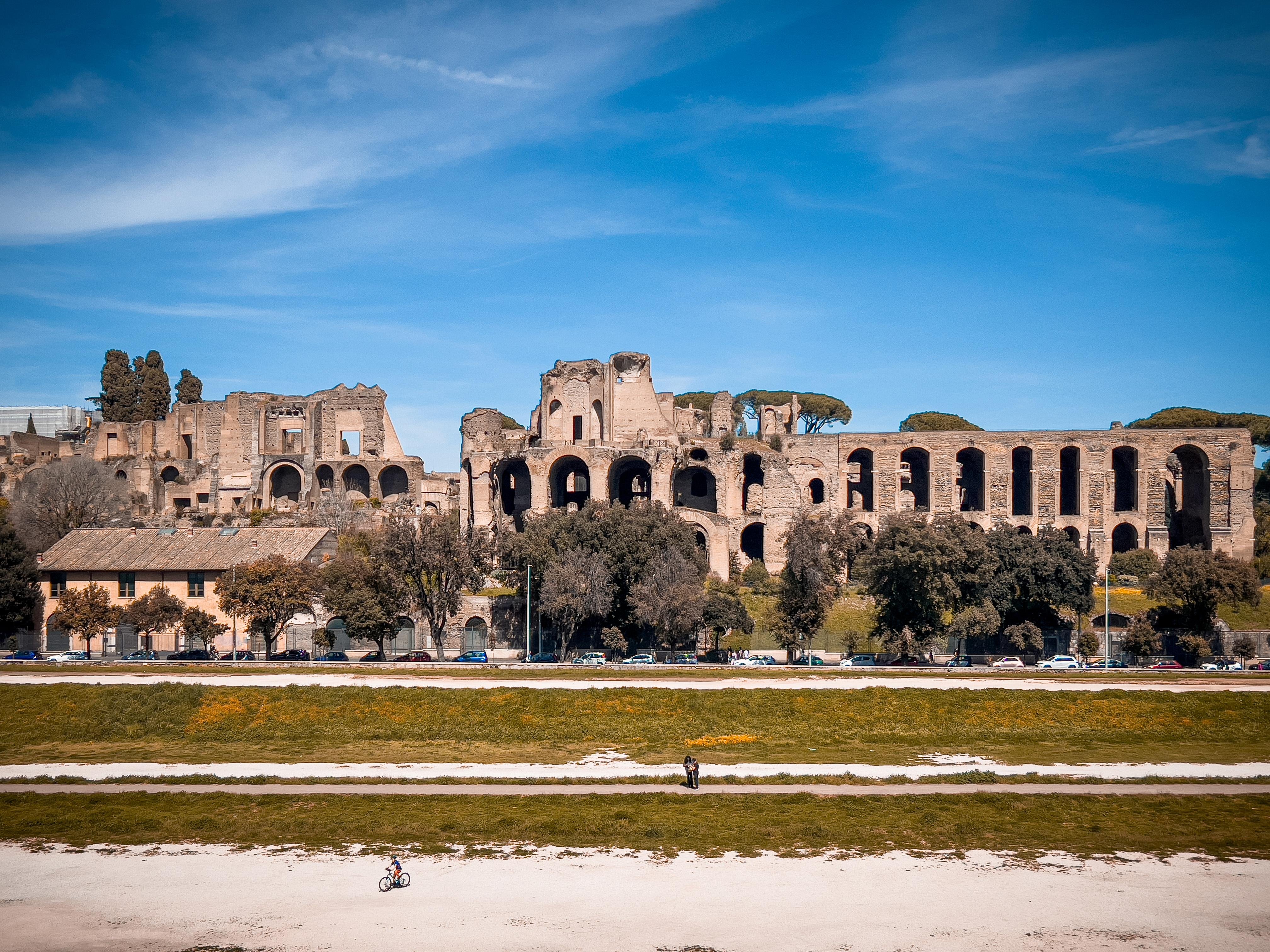Rovine del Palazzo di Domiziano sul Colle Palatino a Roma, Italia. Queste antiche strutture romane sono visibili sotto un cielo azzurro e limpido, con un po' di vegetazione e alberi davanti. C'è un sentiero con una persona che cammina e qualcuno che va in bicicletta in primo piano.