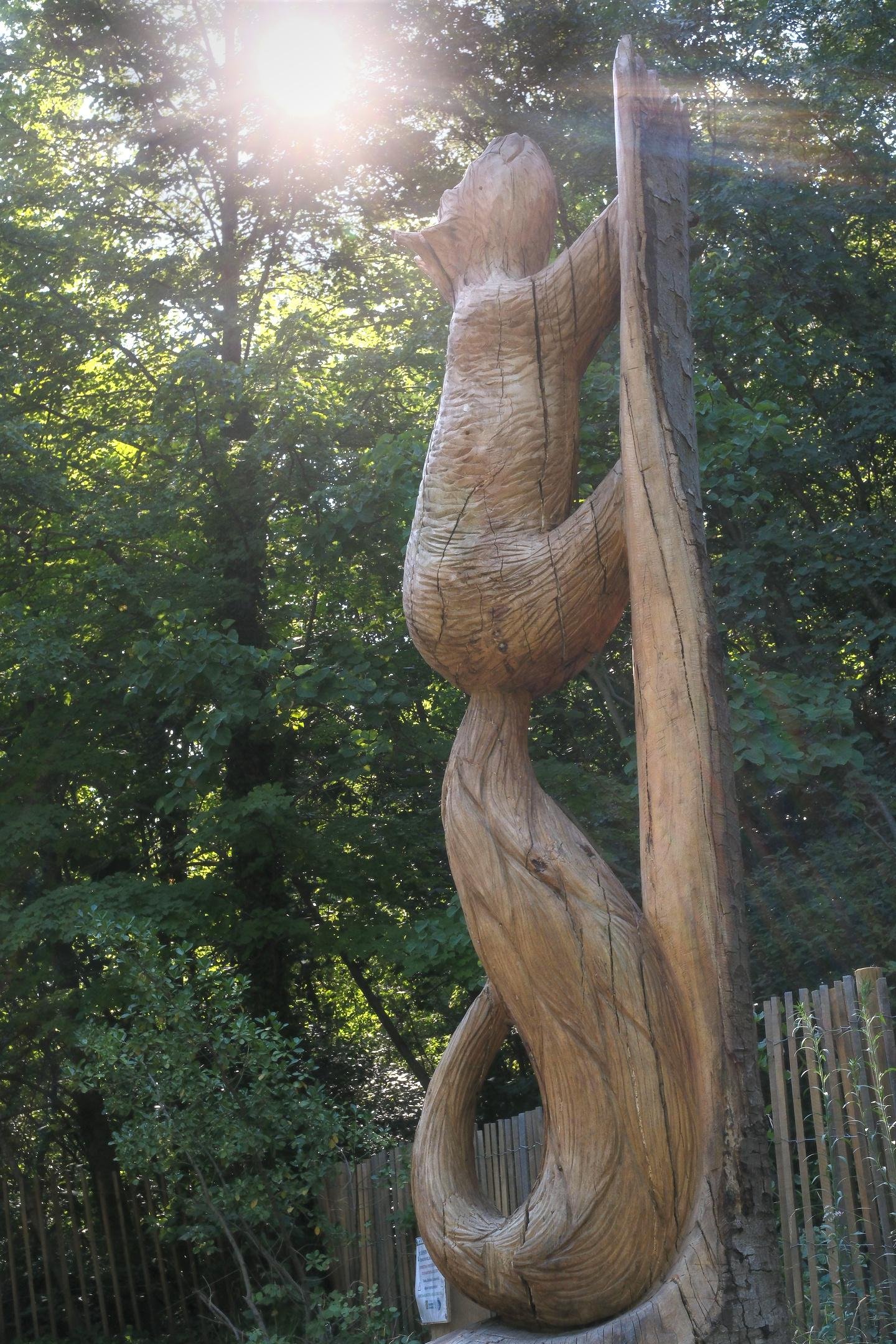 Photo format portrait d’une sculpture géante en bois représentant un écureuil. Le soleil perce à travers le feuillage épais des arbres derrière