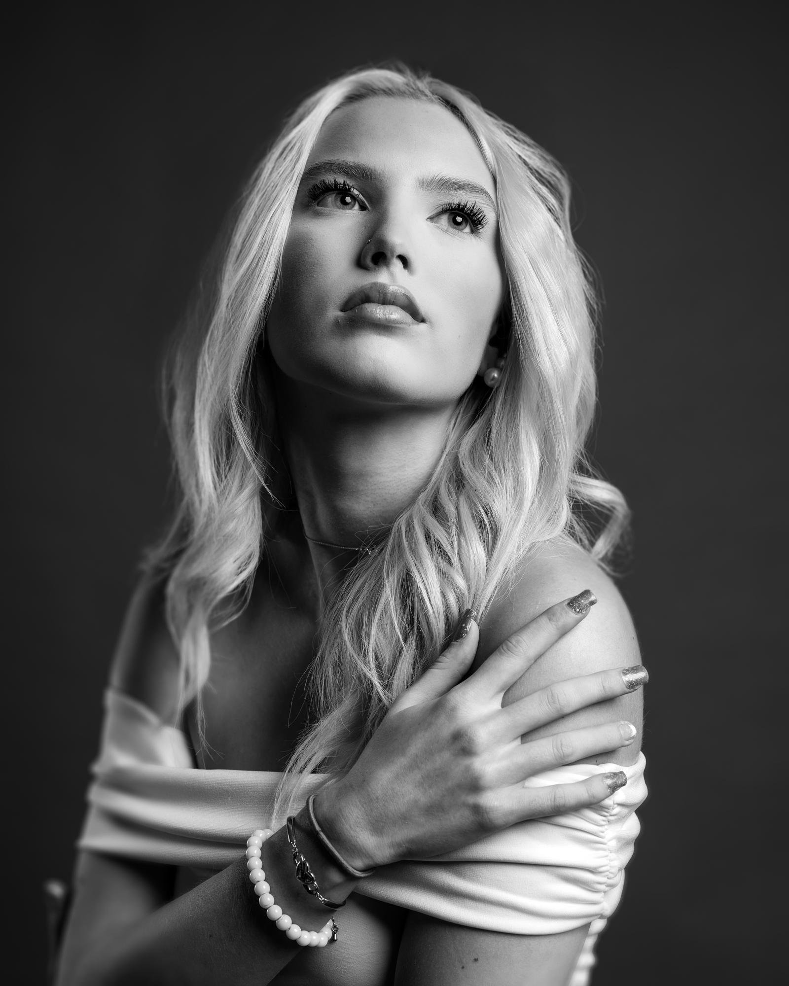 The image is a black and white portrait of a woman with long hair, wearing an off-the-shoulder top. She has her arms crossed, with one hand touching her shoulder, and is looking upwards. The lighting creates a soft and elegant mood. She is wearing bracelets on one wrist.