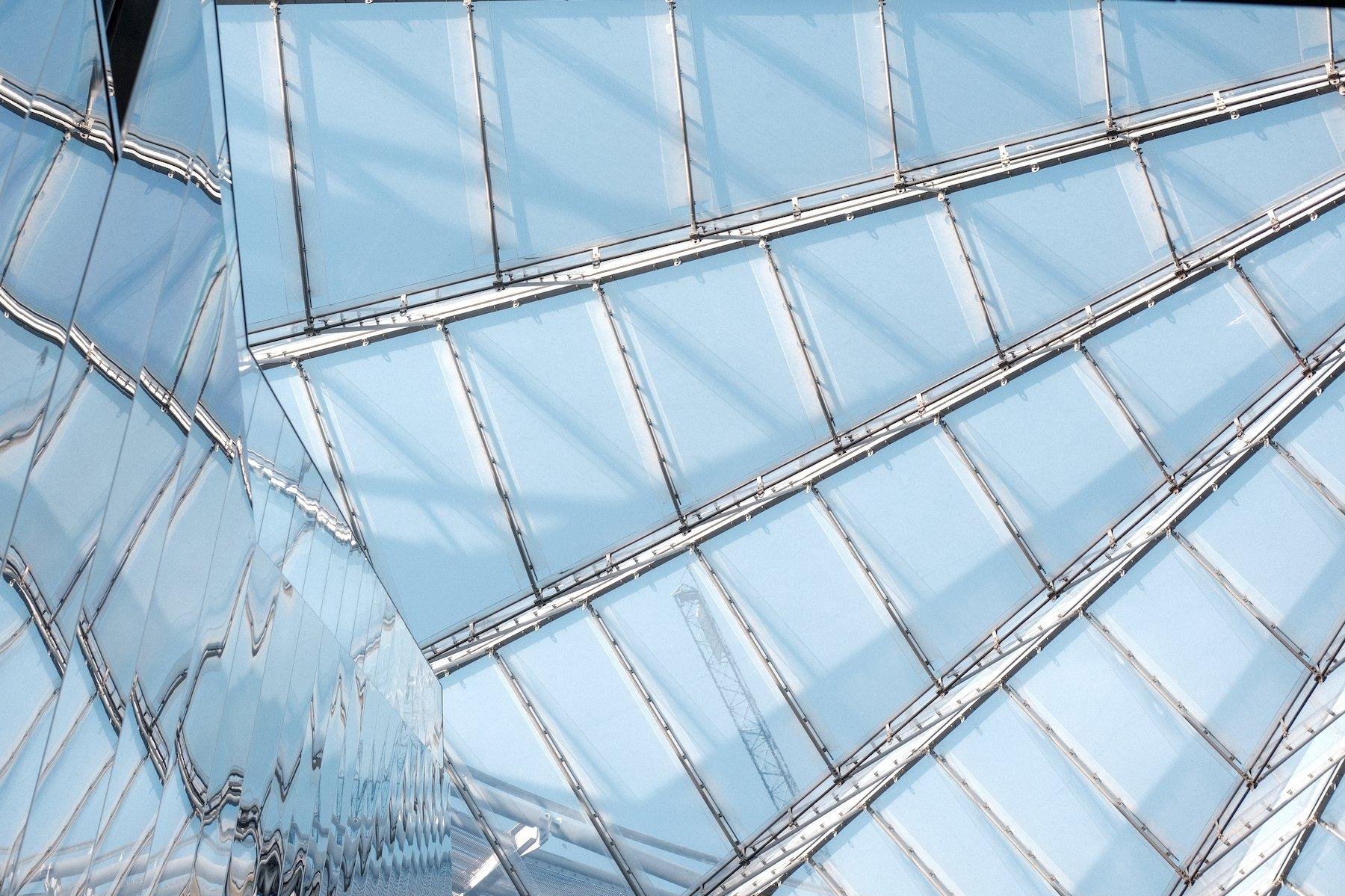 Photo horizontale. Un cadrillage radial du ciel bleu par des vitres d’une verrière gigantesque. Sur la gauche, une surface métallique réfléchissante déforme le cadrillage