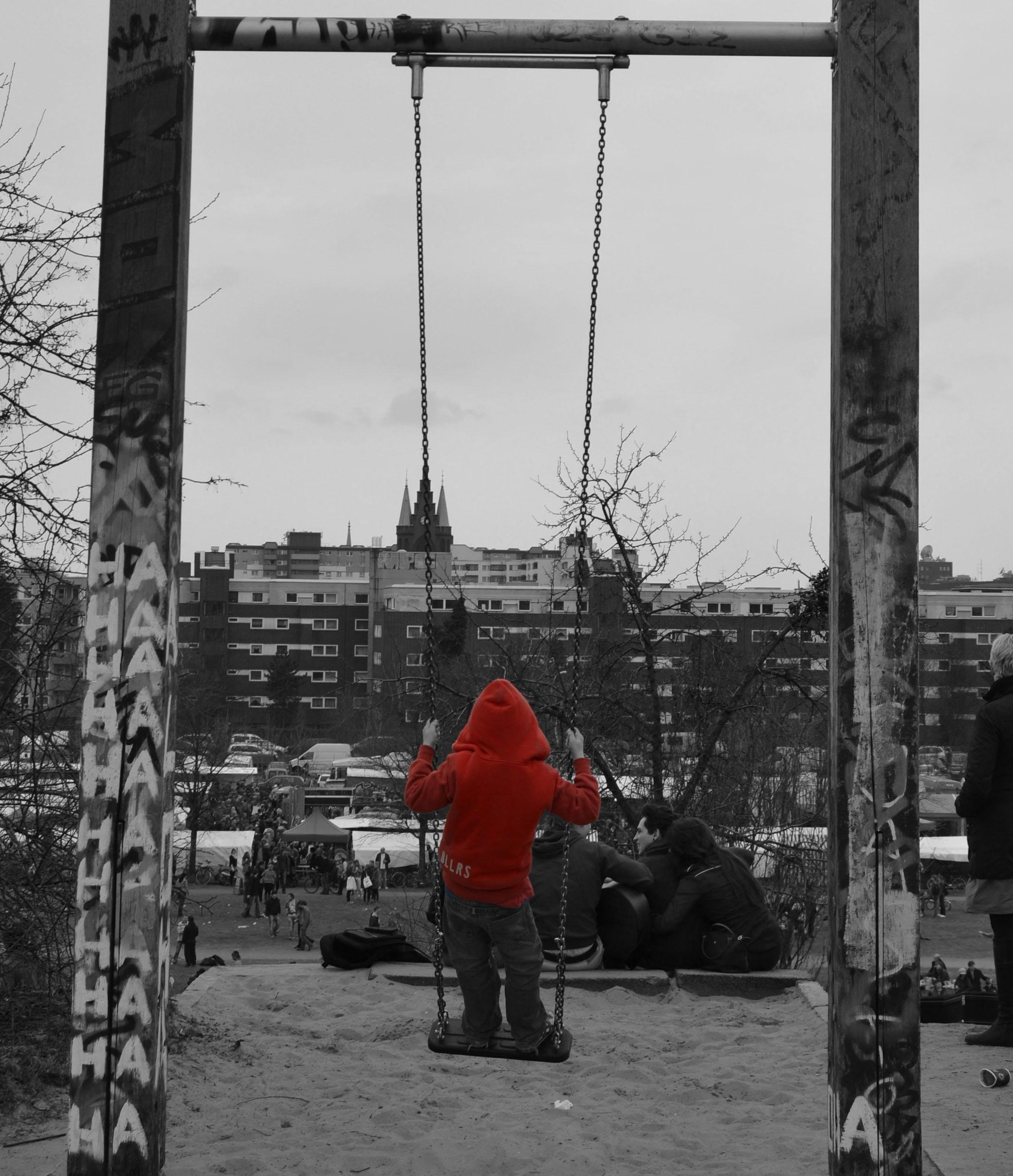 The image shows a child in a red hoodie sitting on a swing. The swing set is covered in graffiti, and there are buildings in the background. The image is mostly black and white, except for the child's red hoodie, which is in color. There are people and a view of a cityscape in the distance.
