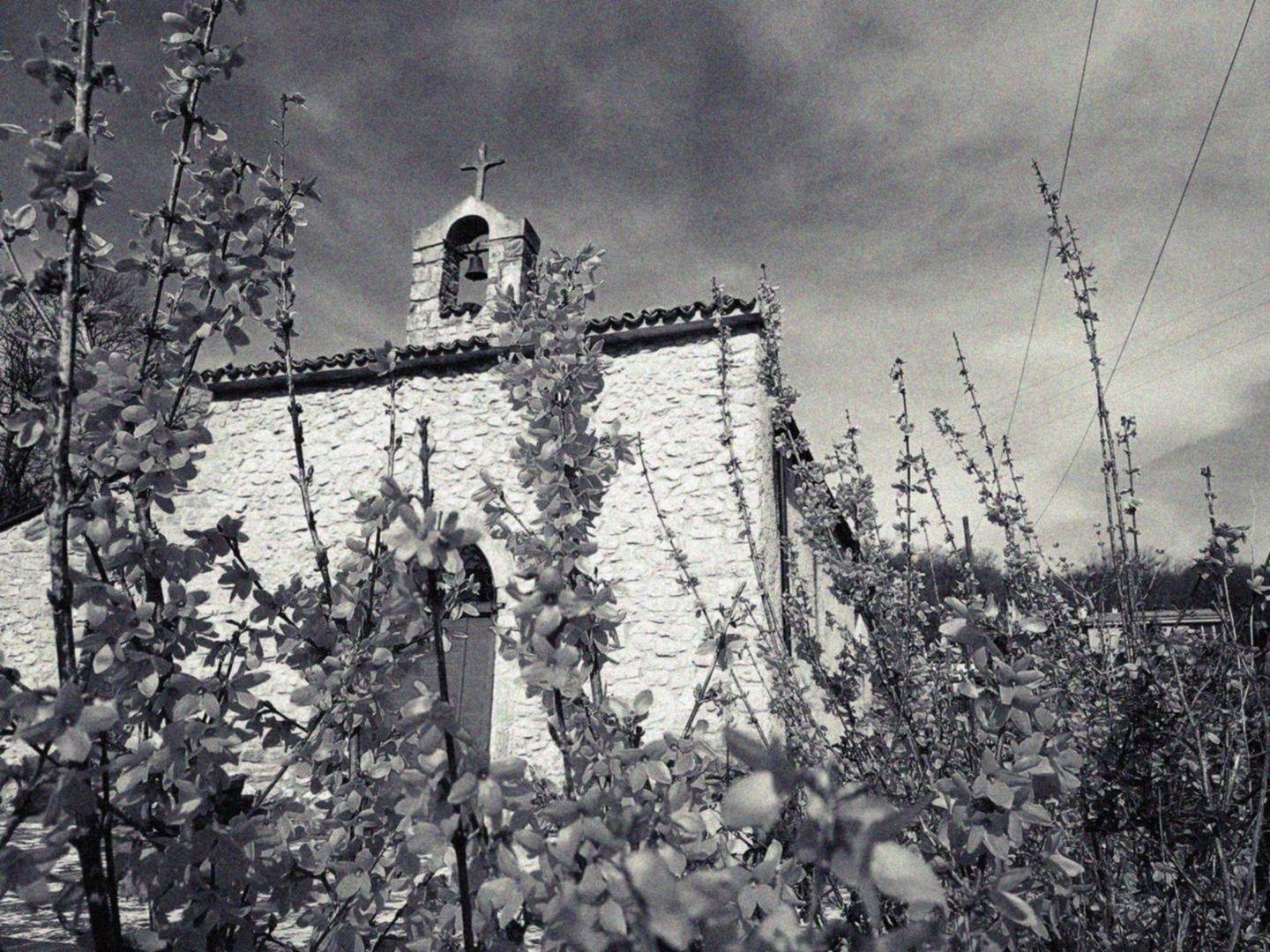 Chiesa di campagnia, con un piccolo campaniele sormontato da una croce, tra il giallo dei fiori in primo piano e il blu del cielo. La foto è in bianco e nero.