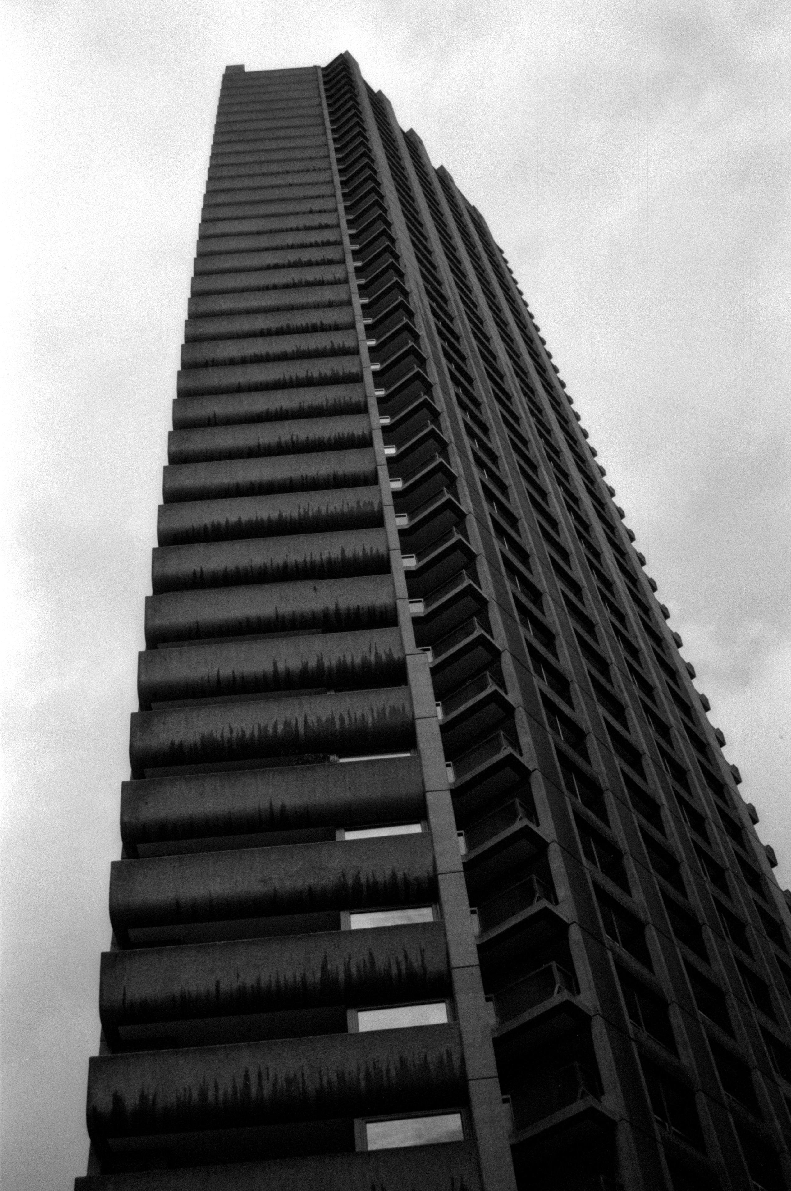 A brutalist-style tower block seen from below, tilting to the left a little. The left-hand side appears to be balconies of some sort and have concrete barriers, curving downwards somewhat for every floor. They have obvious grime near their bottoms, as they have not been cleaned properly. On the right appears to be a different style of balcony with glass barriers, and support pillars moving up the outside of the building.