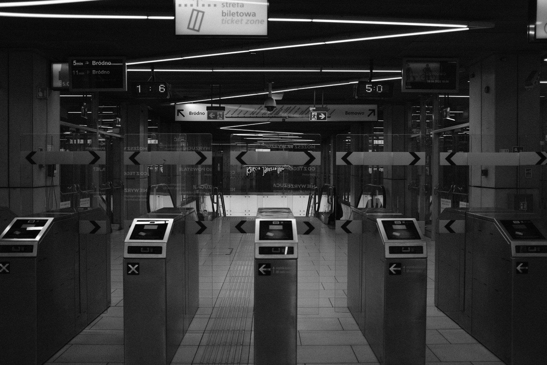 Turnstile gates at a subway station entrance, seen in a black and white photo. Overhead, illuminated signs display directions and platform numbers. The floor is tiled, and ceiling lights form diagonal patterns. Glass barriers with arrow decals are aligned with the gates.