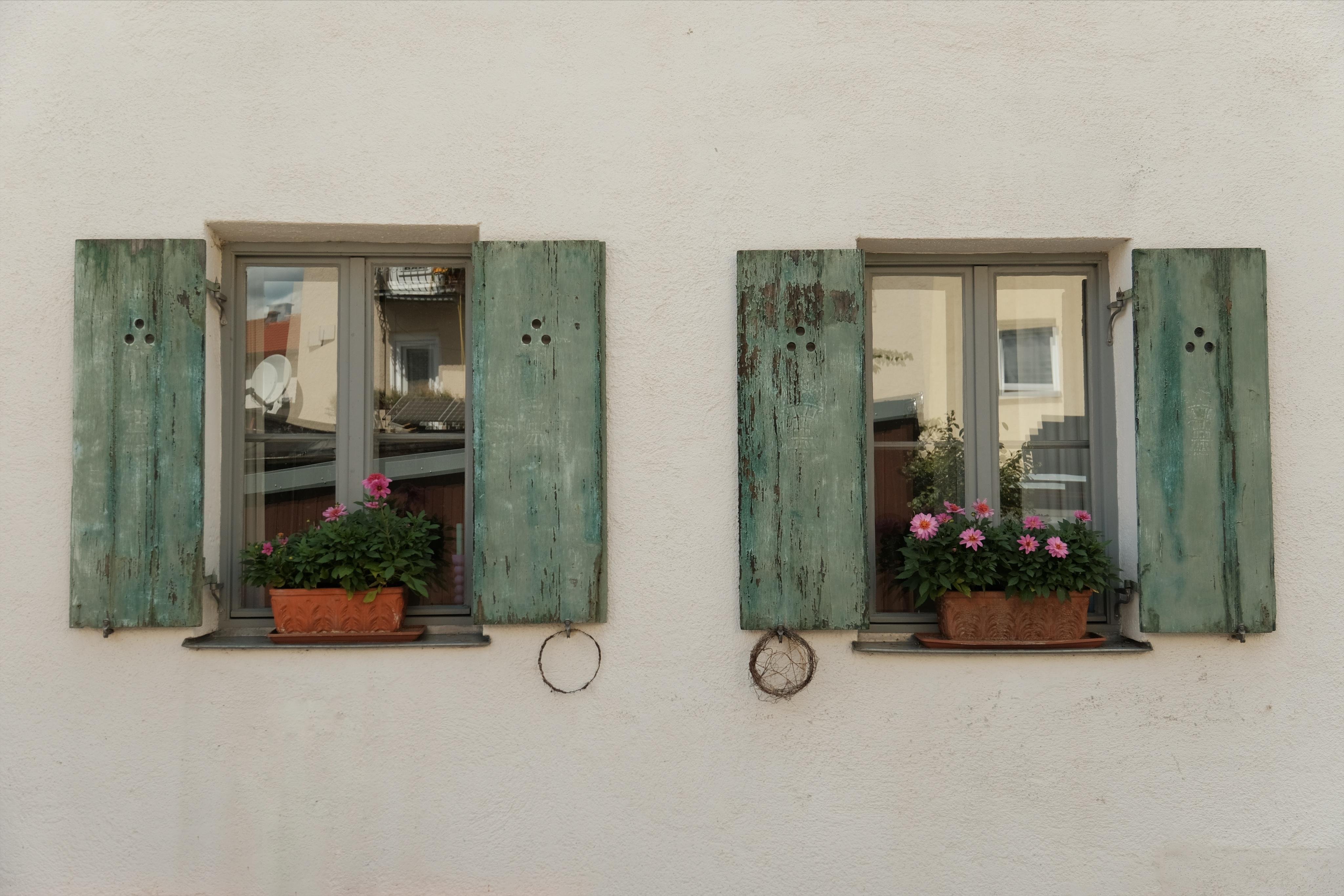Two windows in a white wall with green wooden shutters, all opened symmetrically. Baskets with red and pink flowers on the window sills, decorative elements hanging from the shutters.
