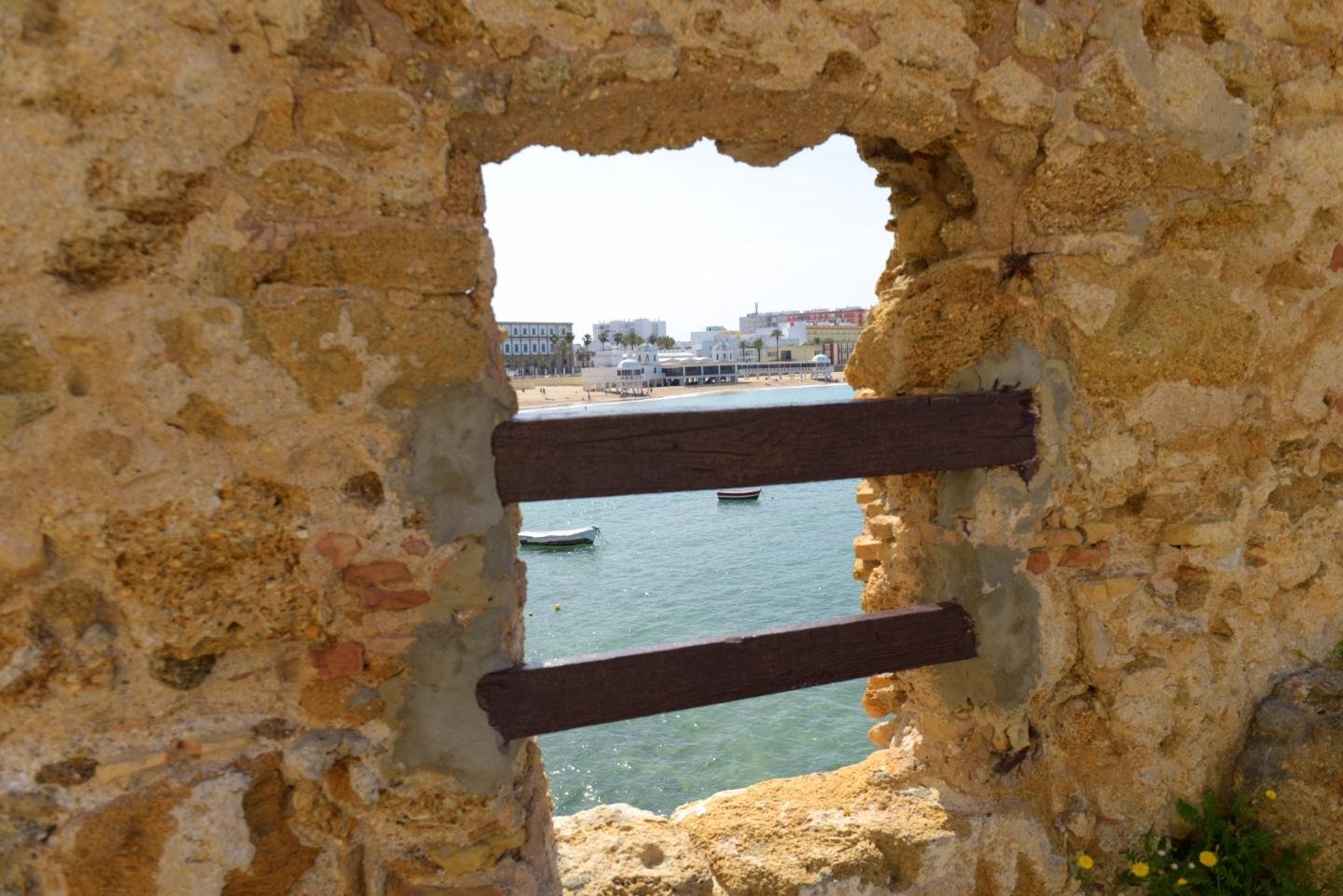 Enmarcado por un paramento de rocas y argamasa desiguales de tonos anaranjados y marrones claros, atravesado por dos listones de madera tomados de cemento gris de mala manera se ve el balneario de la gaditana playa de La Caleta y algún barquillo fondeado.
