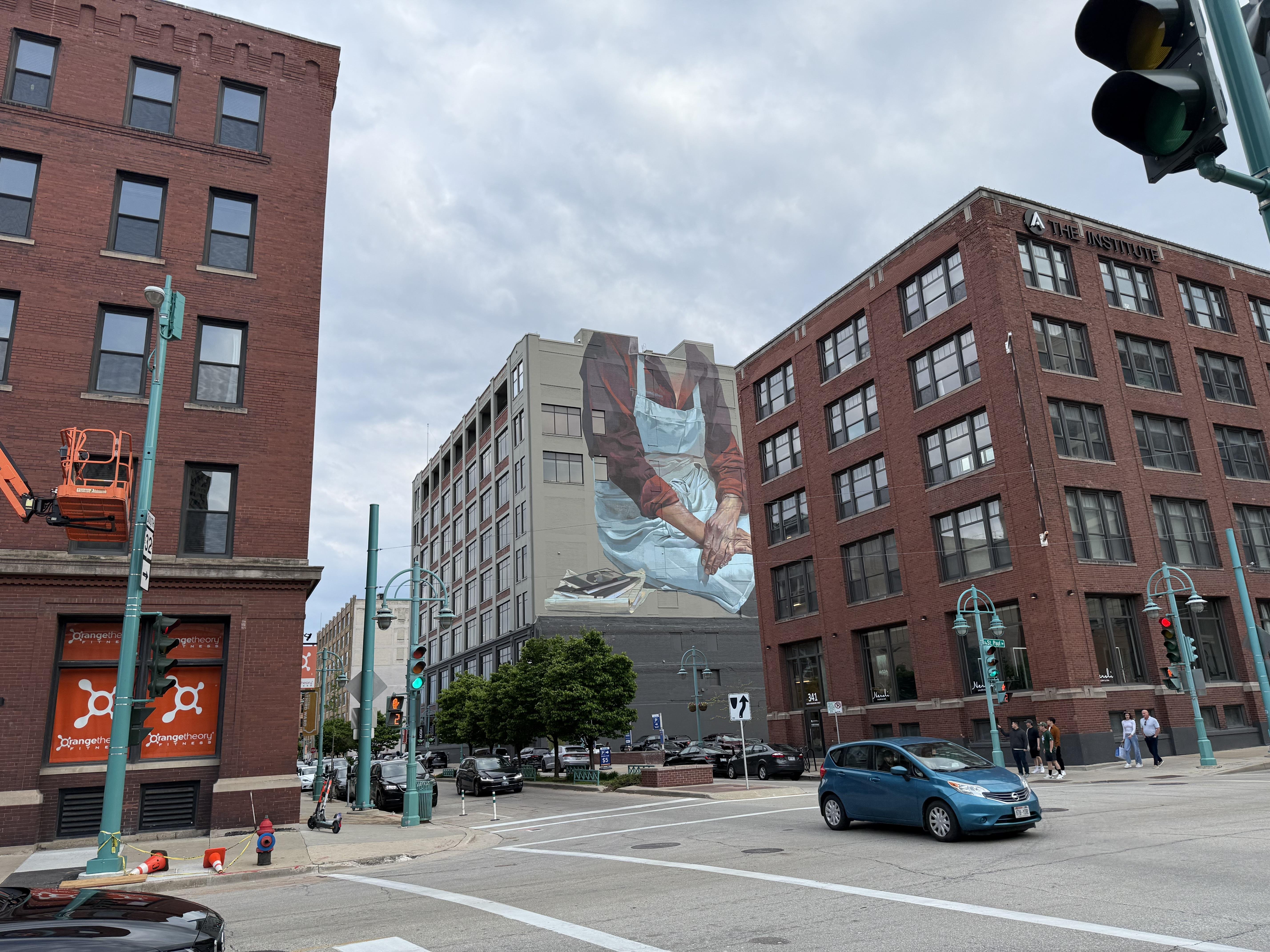 The image shows an urban street scene with several buildings. A prominent mural featuring a person in an apron is visible on the side of one building. There are traffic lights, cars on the road, and pedestrians, with a partly cloudy sky above.