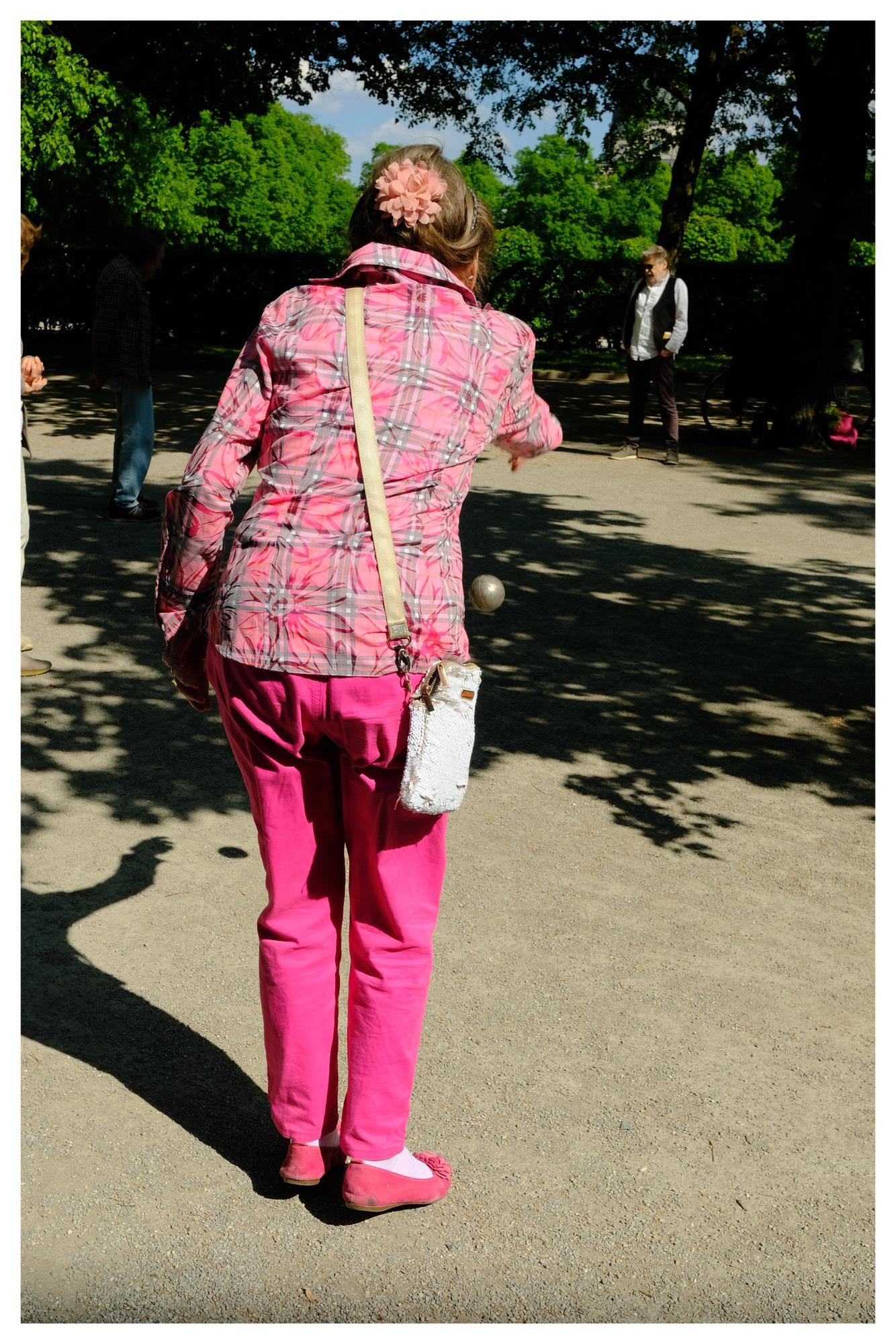 Person seen from behind in a park, wearing a pink plaid jacket and bright pink pants, tossing a metal ball on a gravel pétanque court; trees cast shadows and other players stand in the background.