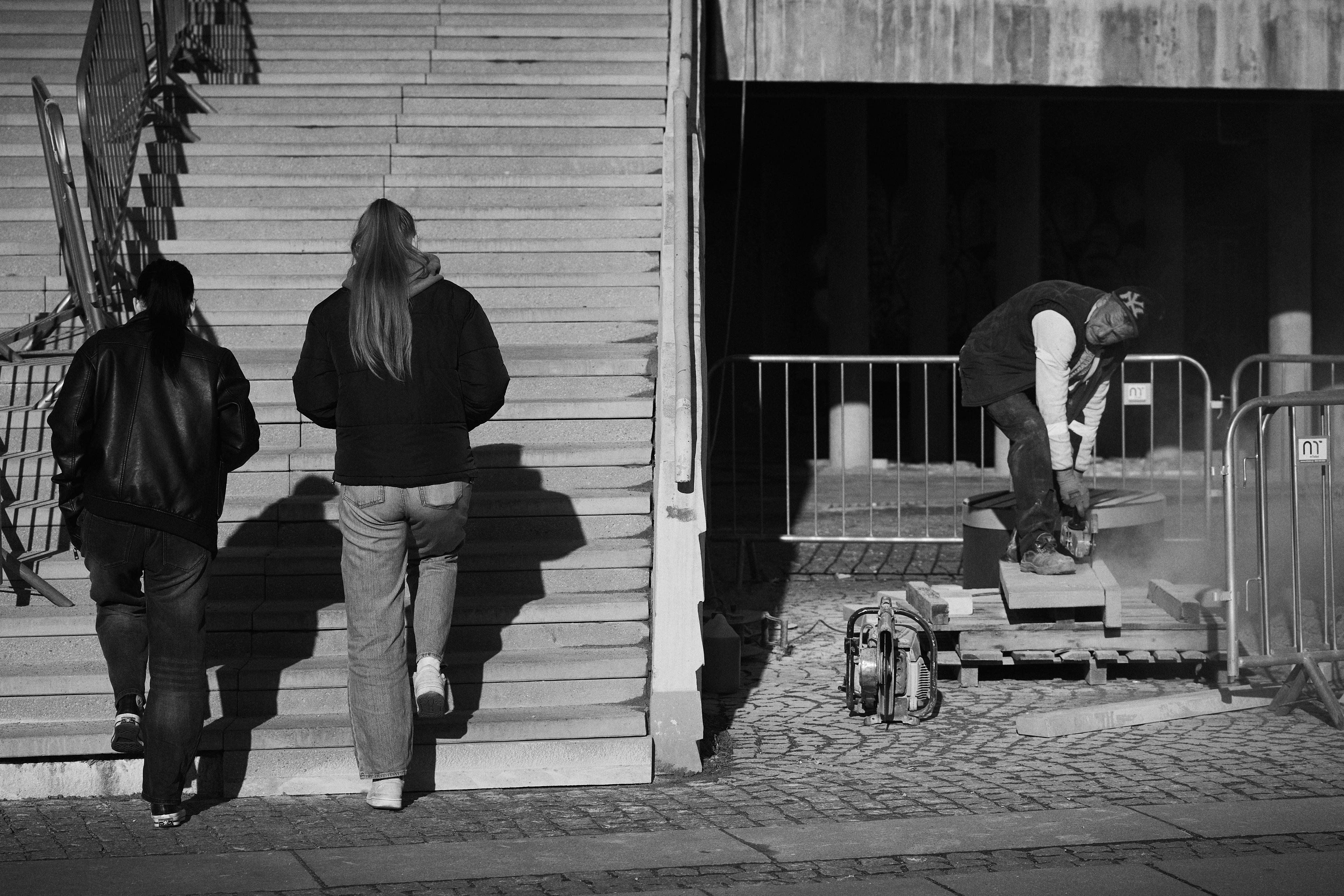 The image shows two people walking up a set of stairs on the left side. On the right, a construction worker is using an electric tool, surrounded by barriers. There is dust being generated by the work, indicating active construction or repair work.