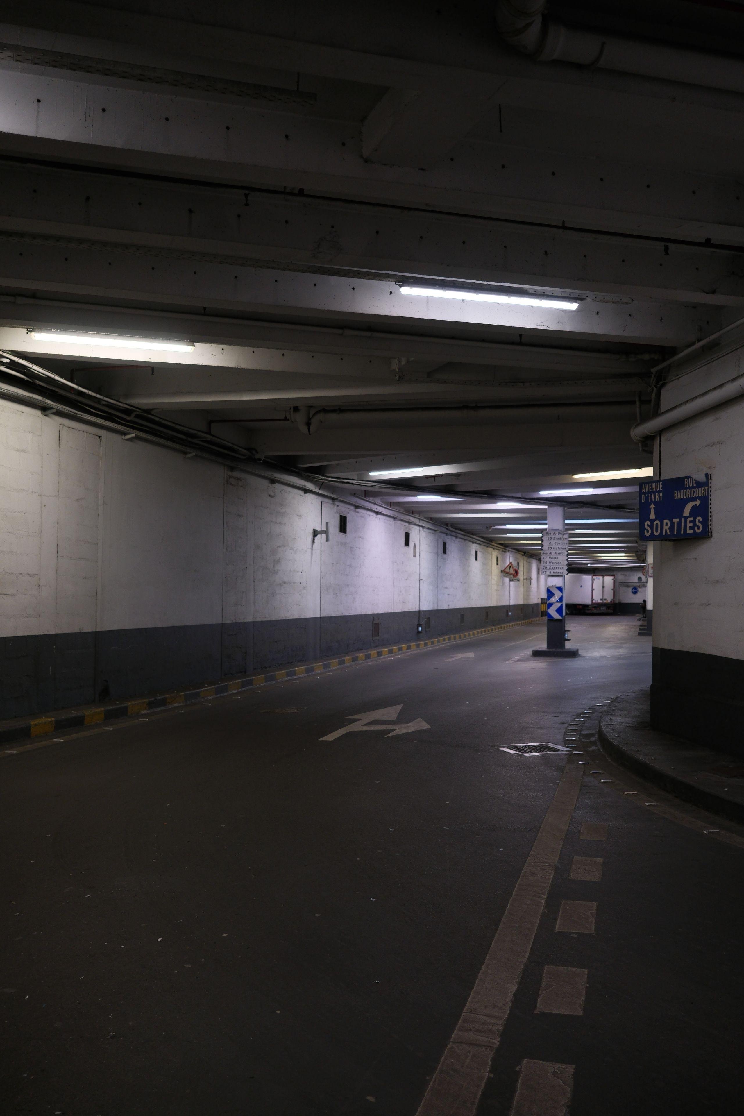 Photo verticale. Une rue souterraine éclairée au néon, avec un marquage au sol indiquant une fourche. Sur un mur à droite, un panneau indiquant les directions