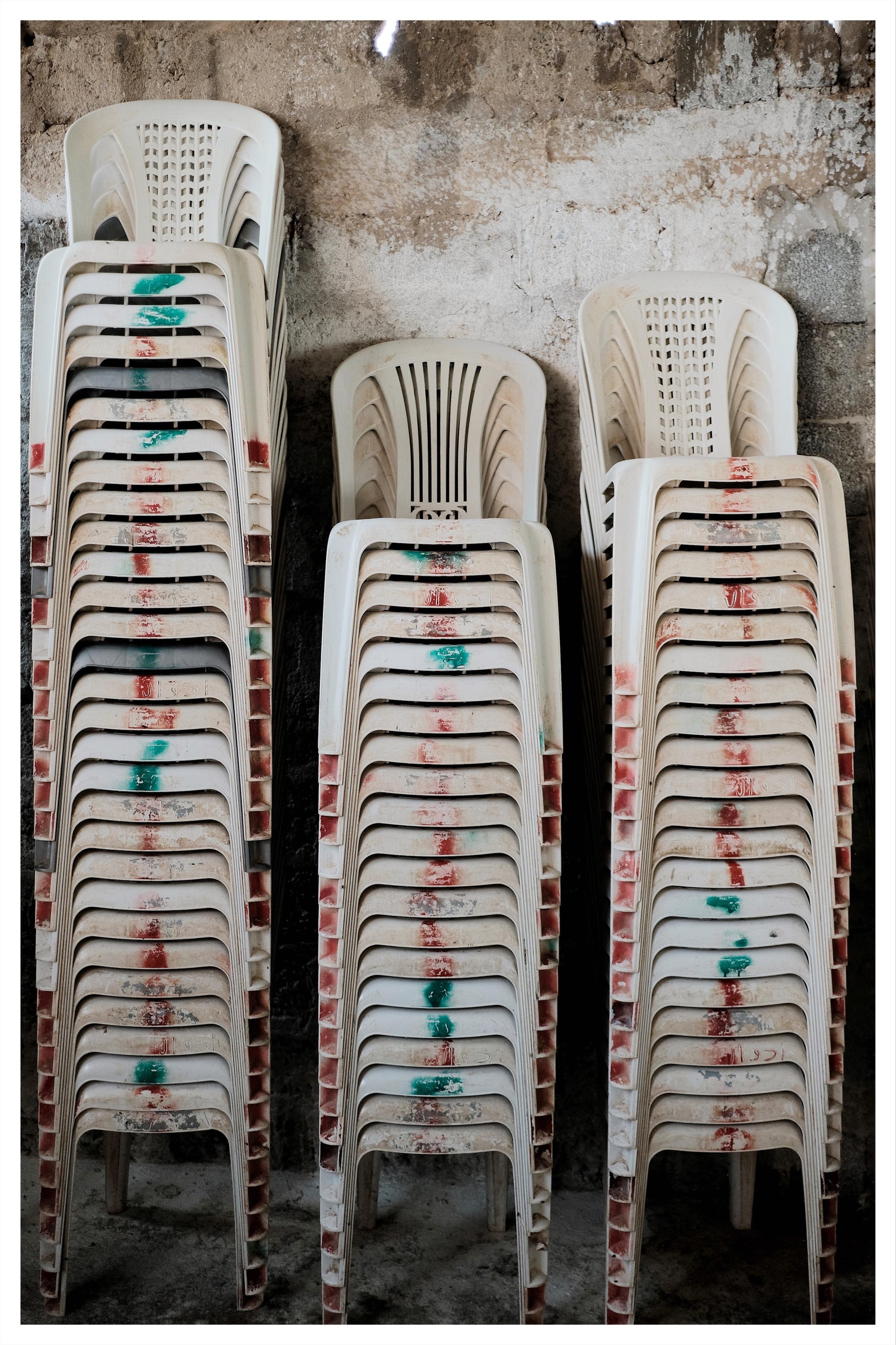 Three tall stacks of white plastic patio chairs with red and green paint marks, arranged against a stained concrete wall on a dusty floor.