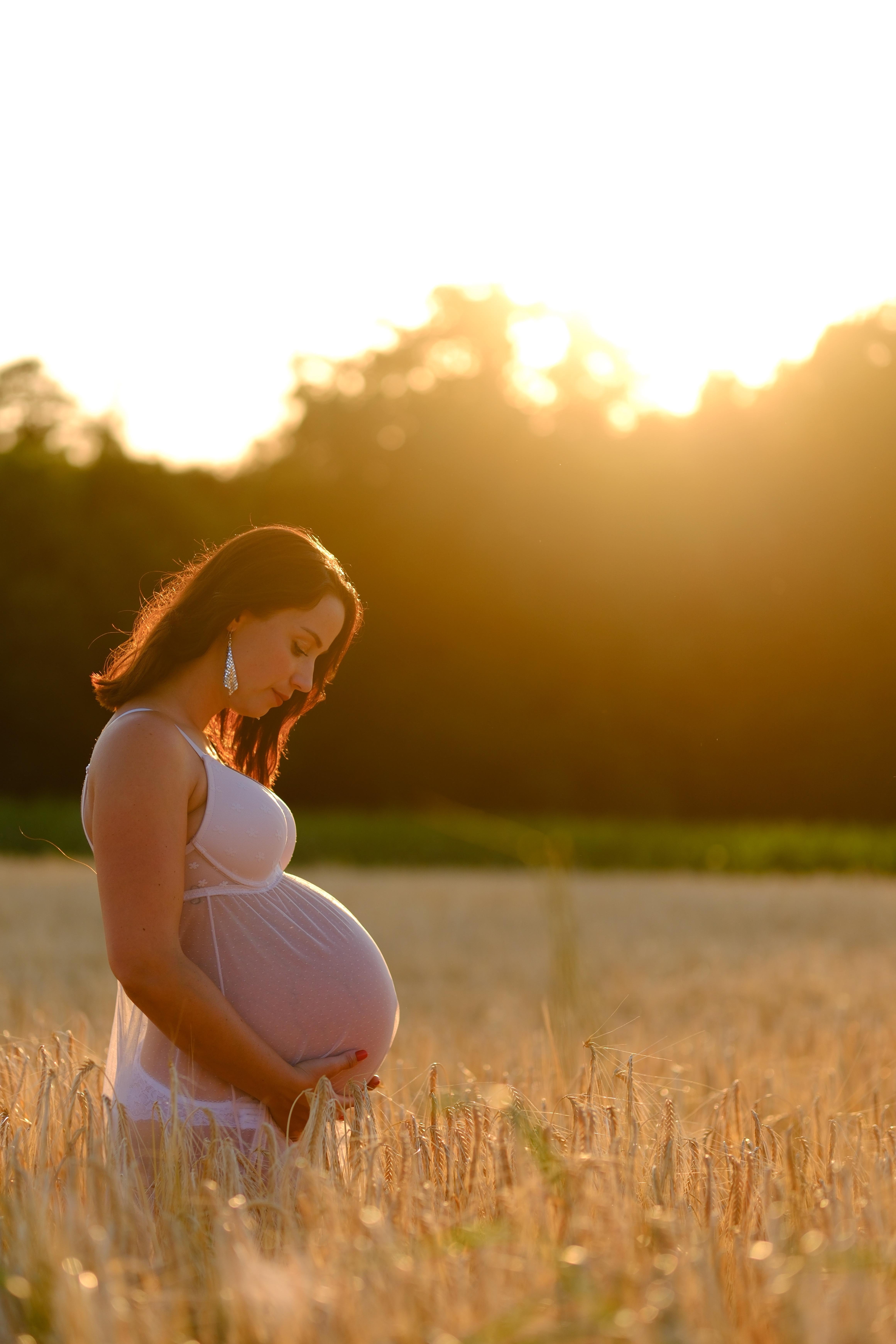 Eine Frau die in eine Feld steht. Ist ist schwanger und hält ihren Bauch. Sie hat ein Kleid an. Im Hintergrund die untergehende Sonne.