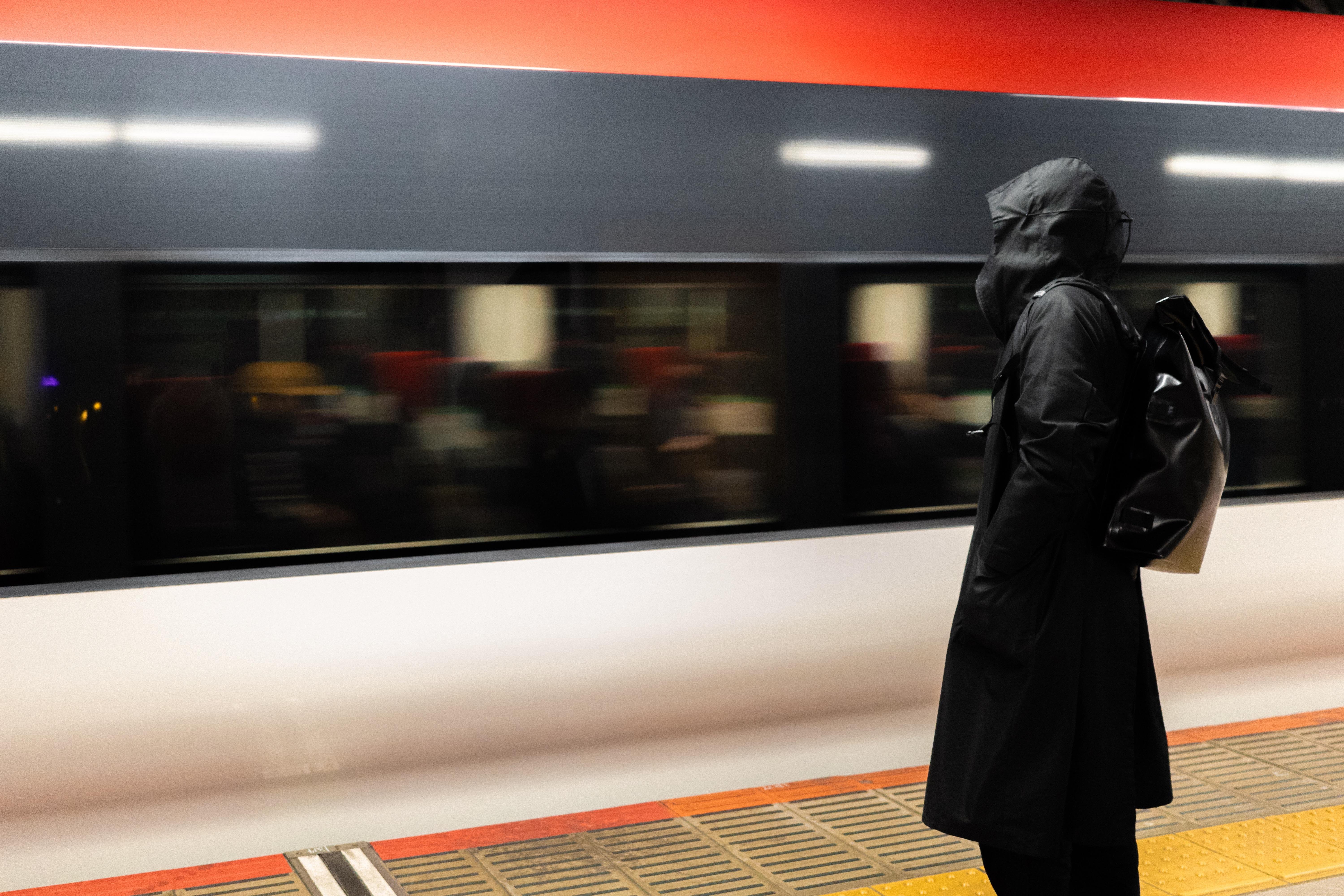 Person in a black hooded raincoat standing on a train platform, facing a speeding train with a blurred motion. The train has a horizontal red stripe near the roof and illuminated windows. The platform edge is marked with yellow and red safety markings.