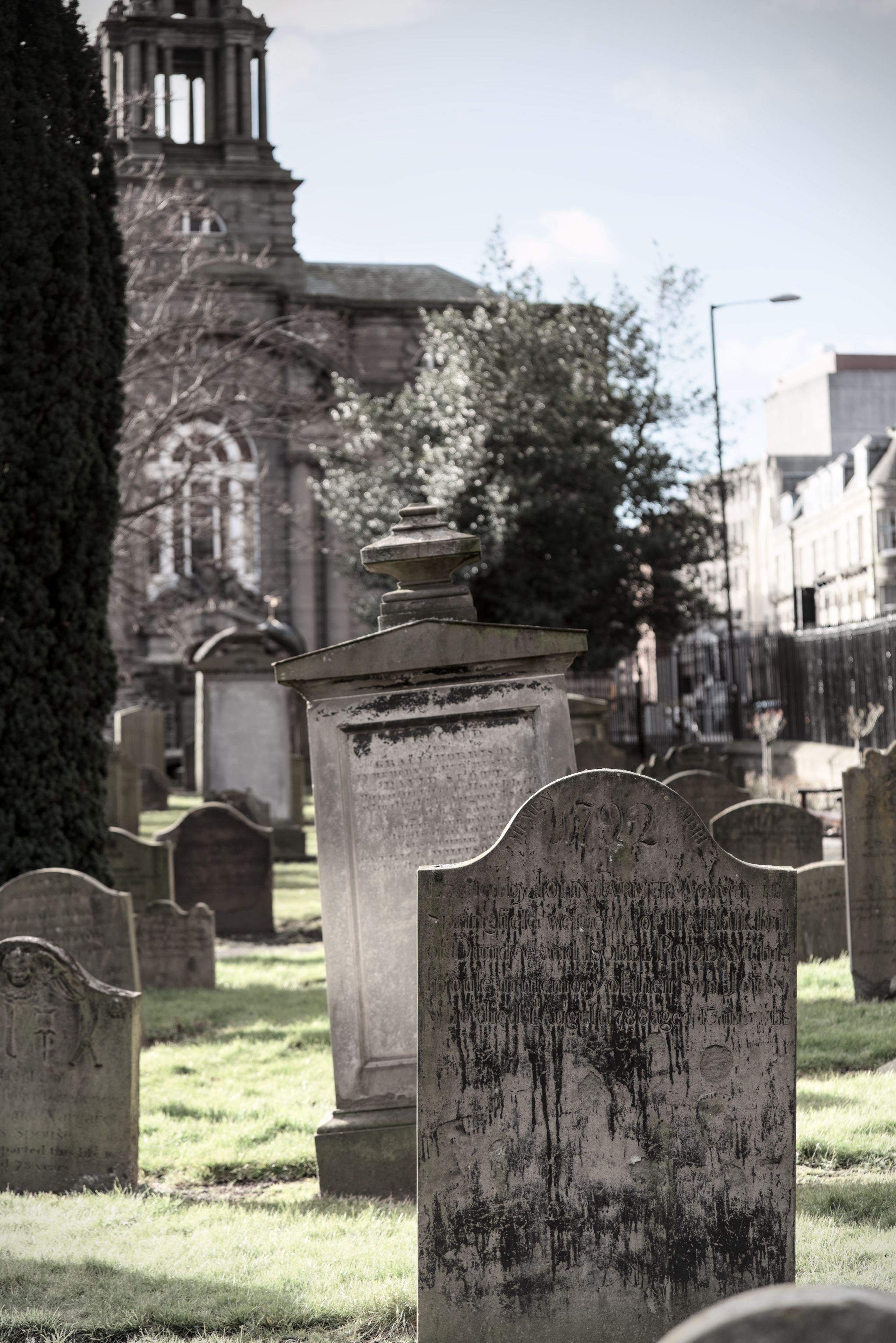 Photo format portrait aux couleurs ternes, montrant de très vieilles pierres tombales (on peut lire 1782 sur l’une d’elle). Celle au premier plan est pleine de coulures sombres et celle juste derrière est légèrement penchée sur la gauche, d’autres tombes plus petites sont un peu partout à droite comme à gauche