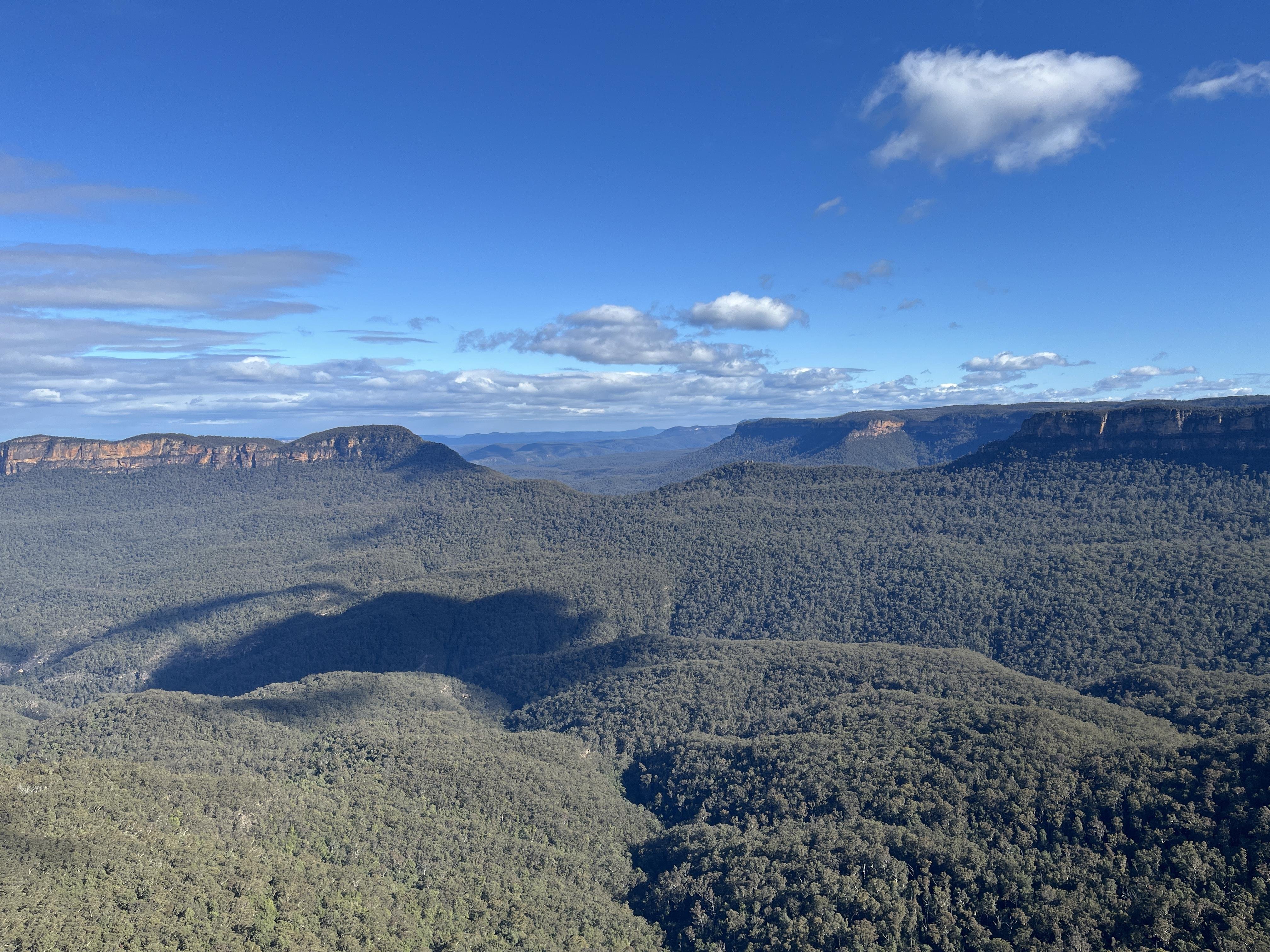 Vast landscape of rolling green forested hills under a clear blue sky. A line of rugged cliffs is visible in the distance, with scattered white clouds above. Shadows from the clouds create darker patches on the hills below.