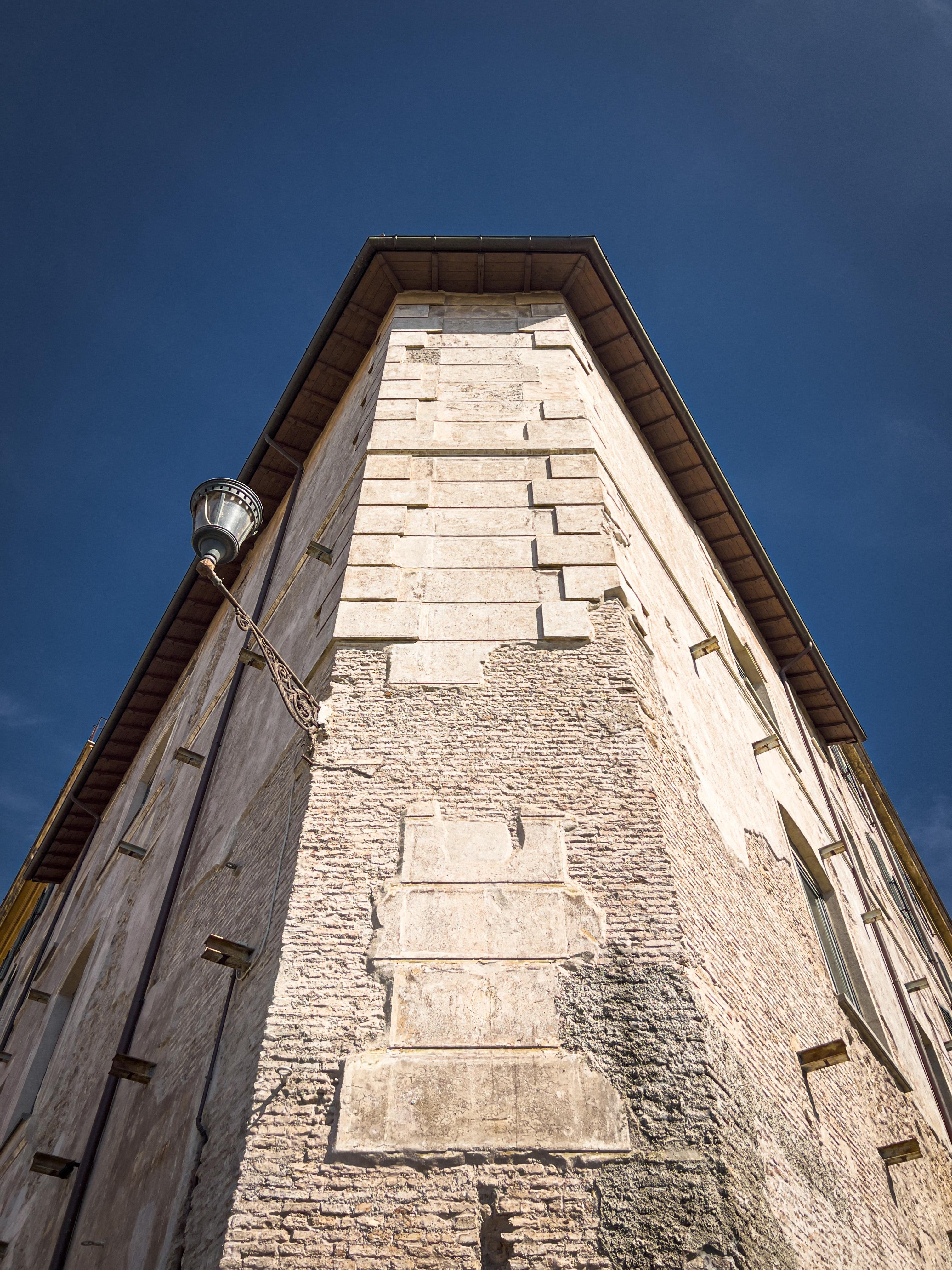 Vista ravvicinata della facciata di un edificio storico da un'angolazione bassa. La struttura sembra essere fatta di pietra o mattoni e presenta elementi architettonici classici. Sulla sinistra c'è un lampione ornato, e il cielo è limpido e blu.