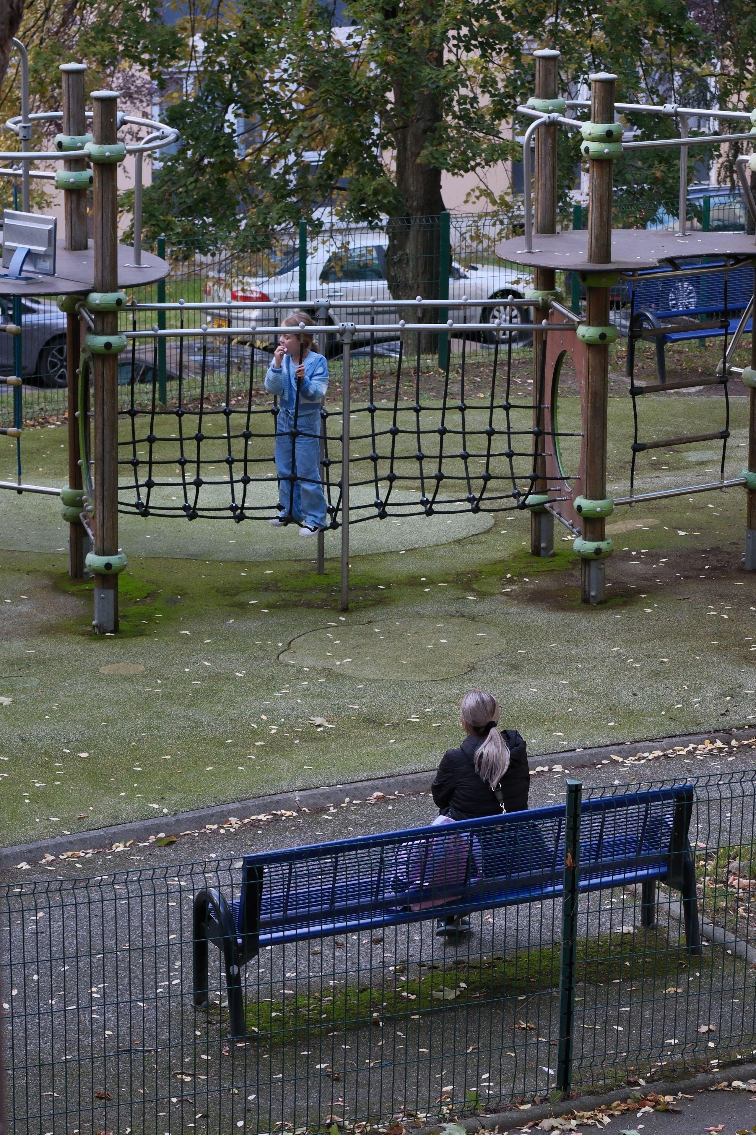 Photo verticale. Une femme assise sur un blanc bleu surveille son enfant qui joue sur une aire de jeux