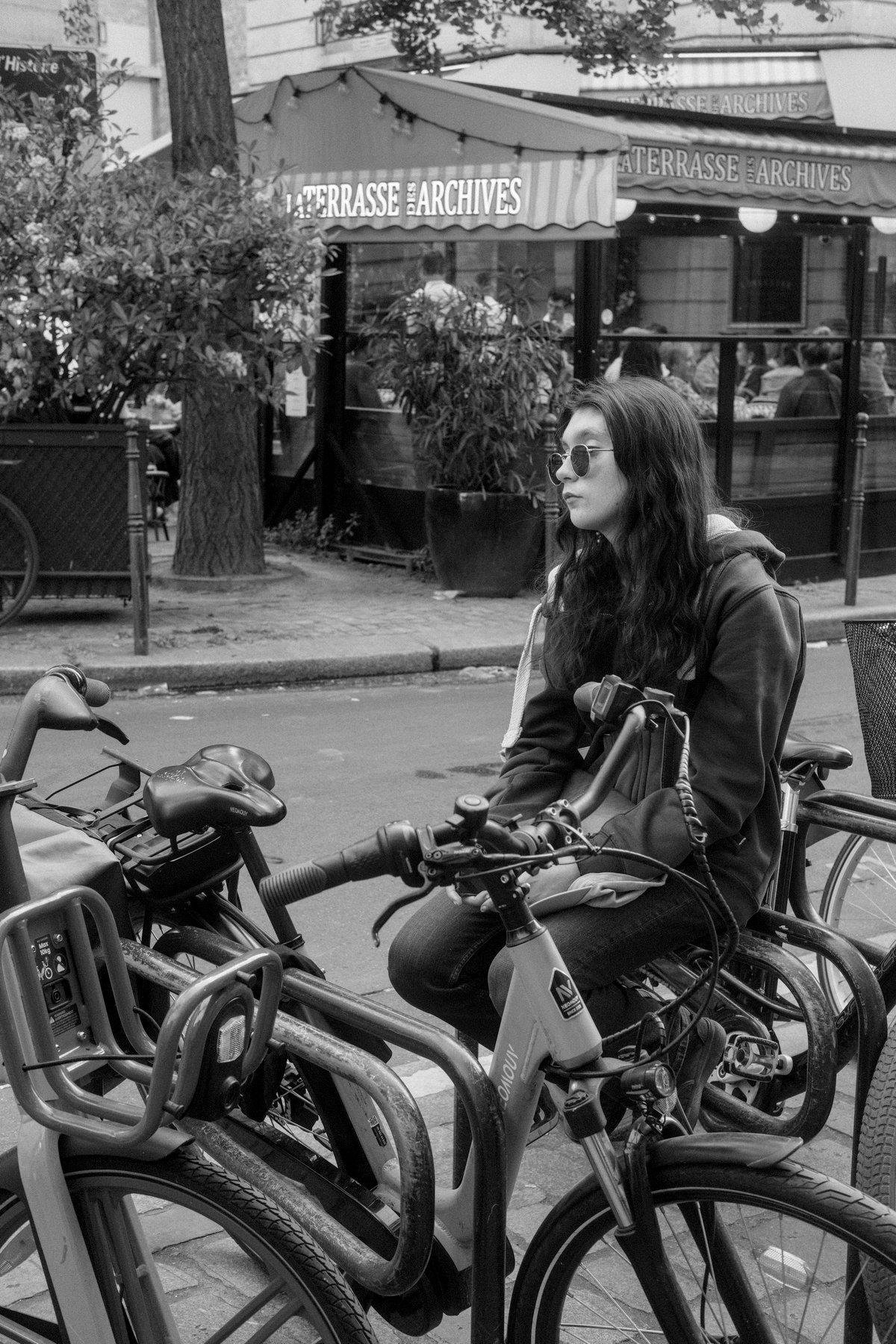 Photo verticale en noir et blanc. Une jeune femme aux cheveux long et sombres, légèrement ondulés, en hoodie, assise sur une place d’attache pour vélo regarde dans le vide derrière ses lunettes de soleil rondes