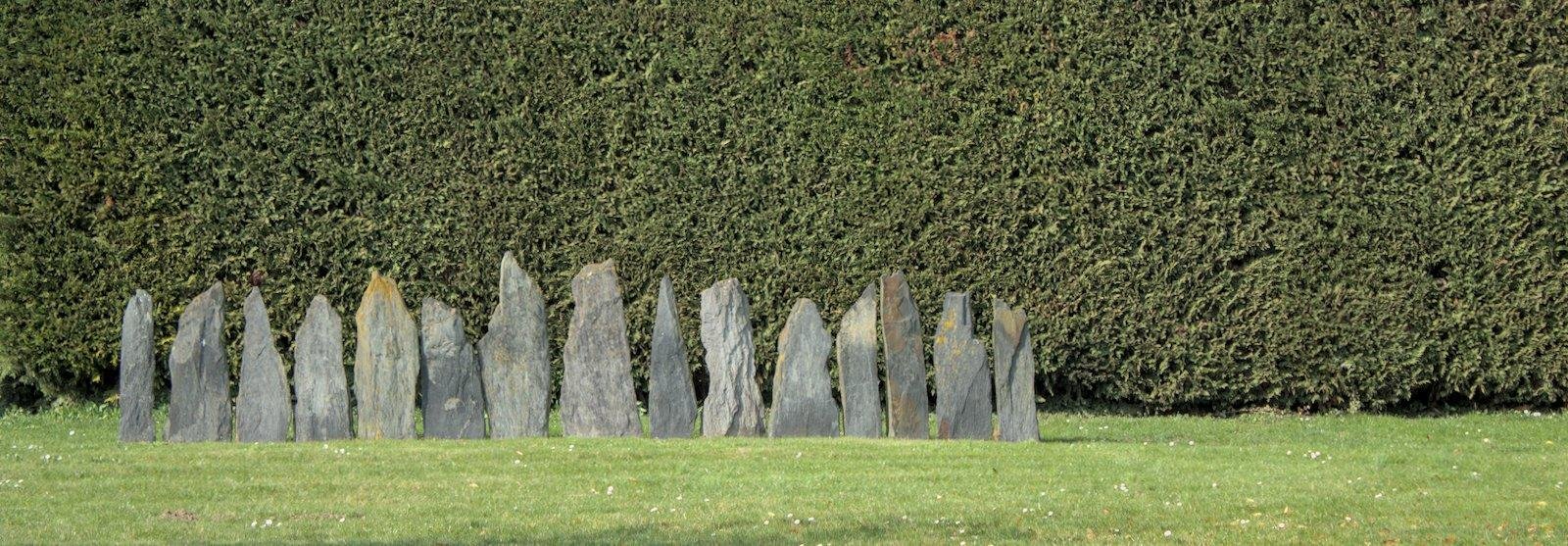 Photo horizontale format widescreen de 15 stèles en ardoise claire de différentes hauteurs, plantées côte à côte sur de la pelouse, devant une haie bien taillée