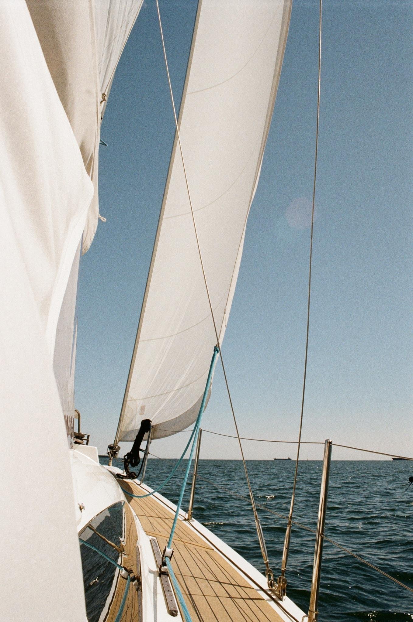 View along the deck of a sailboat with white sails and rigging lines against a clear blue sky, open sea on the right, and a distant ship near the horizon.
