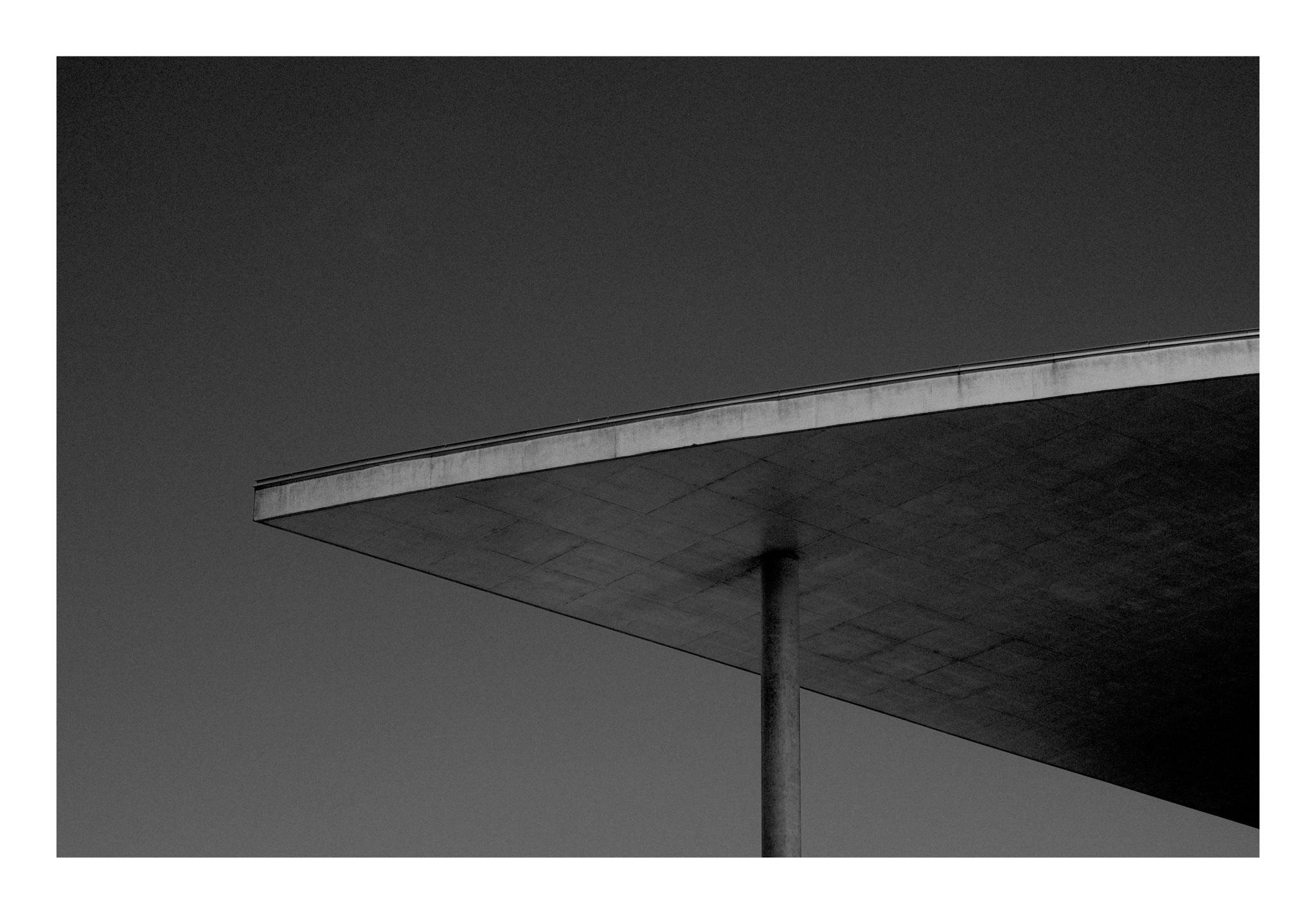 Black-and-white photo of a large flat roof canopy extending diagonally across the upper right, supported by a single thin pole against an empty sky.
