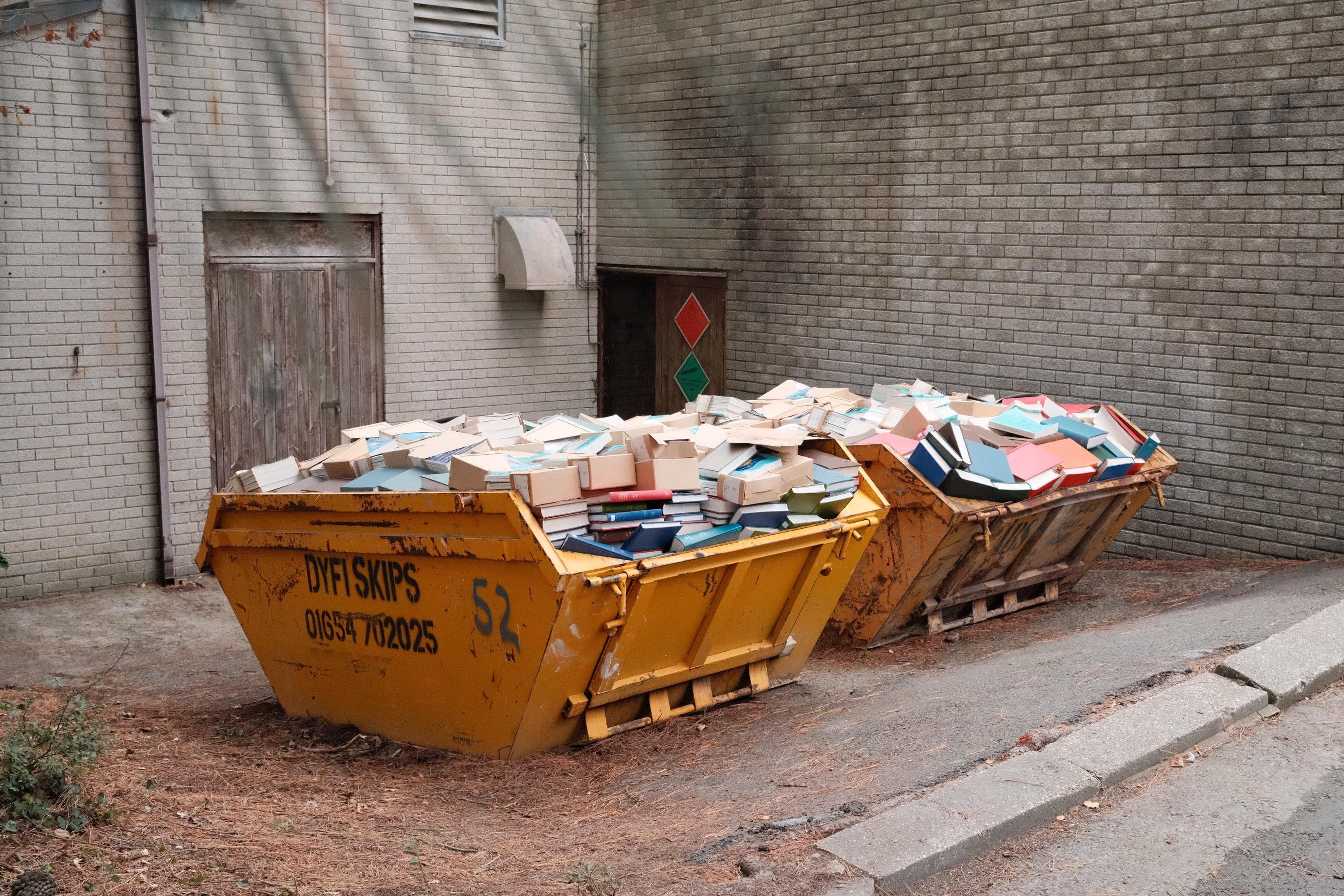 Two yellow metal skips overflowing with academic textbooks and binders in an alley beside a grey brick university building, with a sloped curb and a closed wooden door in the background.