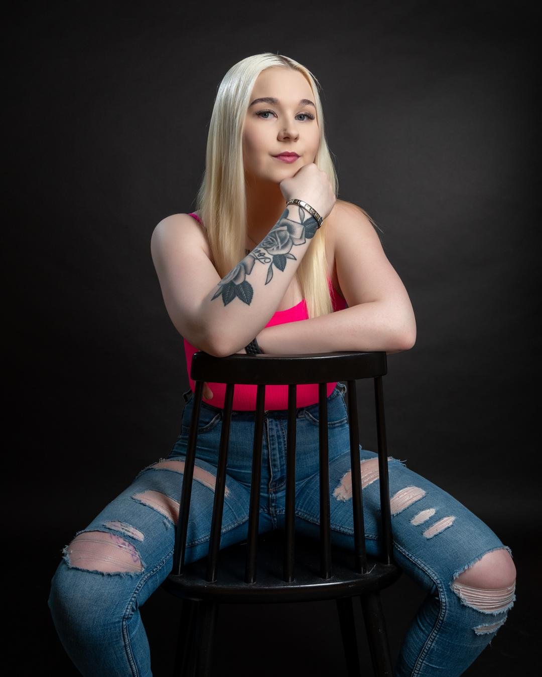 The image shows a person with long blonde hair sitting on a black chair. They are wearing a bright pink top and ripped blue jeans. They have a prominent tattoo on their forearm and are posing with their chin resting on their hand. The background is dark.