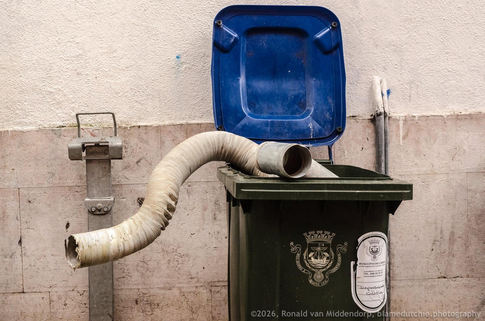 Green wheeled trash bin with its blue lid open against a wall, with a large flexible white hose draped into the bin and hanging out over the left side; metal fixture on the left and small pipes on the right.