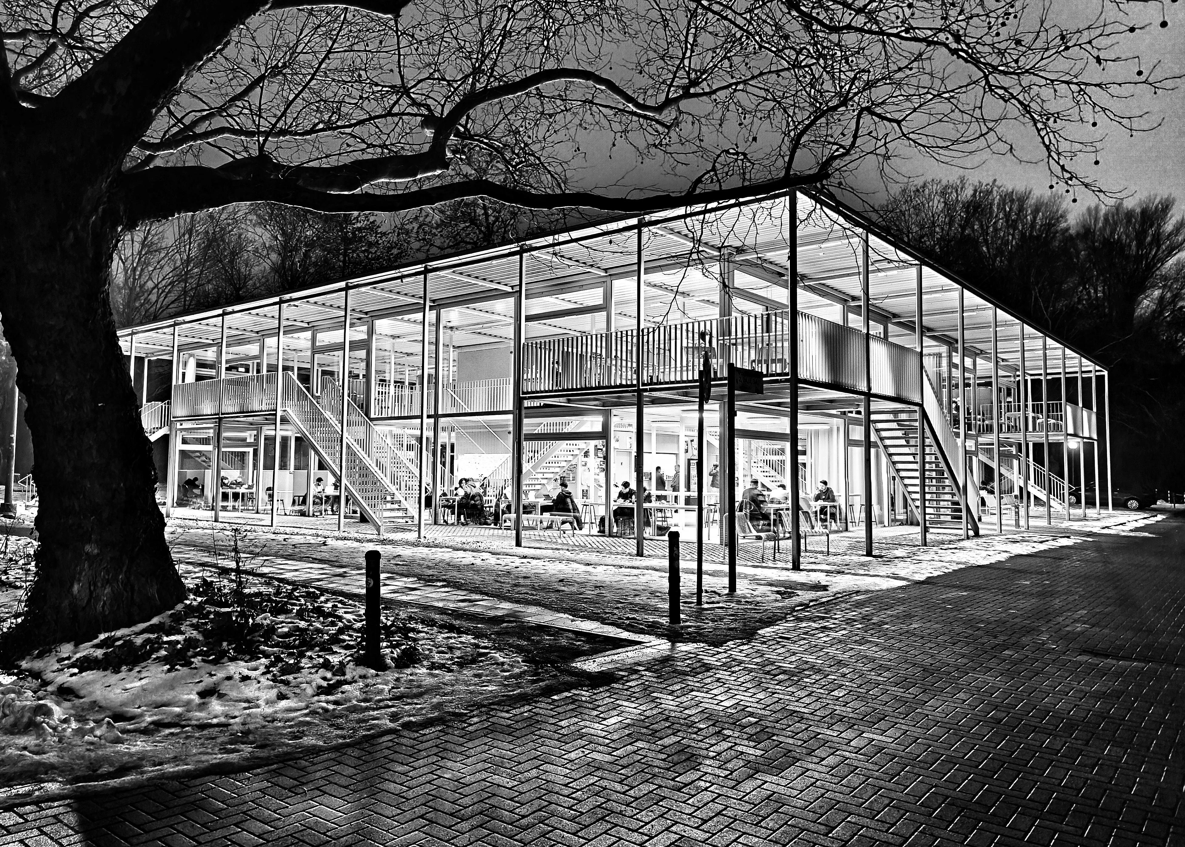 Two-story glass-and-metal pavilion at night with bright interior lights, exterior staircases and wraparound balcony; leafless tree branches frame the top and left, and a brick path with patches of snow leads past the building where seated people are visible inside. 
### 
Ein zweistöckiger Pavillon aus Glas und Metall bei Nacht mit heller Innenbeleuchtung, Außentreppen und umlaufendem Balkon; blattlose Äste rahmen die Oberseite und die linke Seite ein, und ein Backsteinweg mit Schneeflecken führt an dem Gebäude vorbei, in dem sitzende Personen zu sehen sind. 
