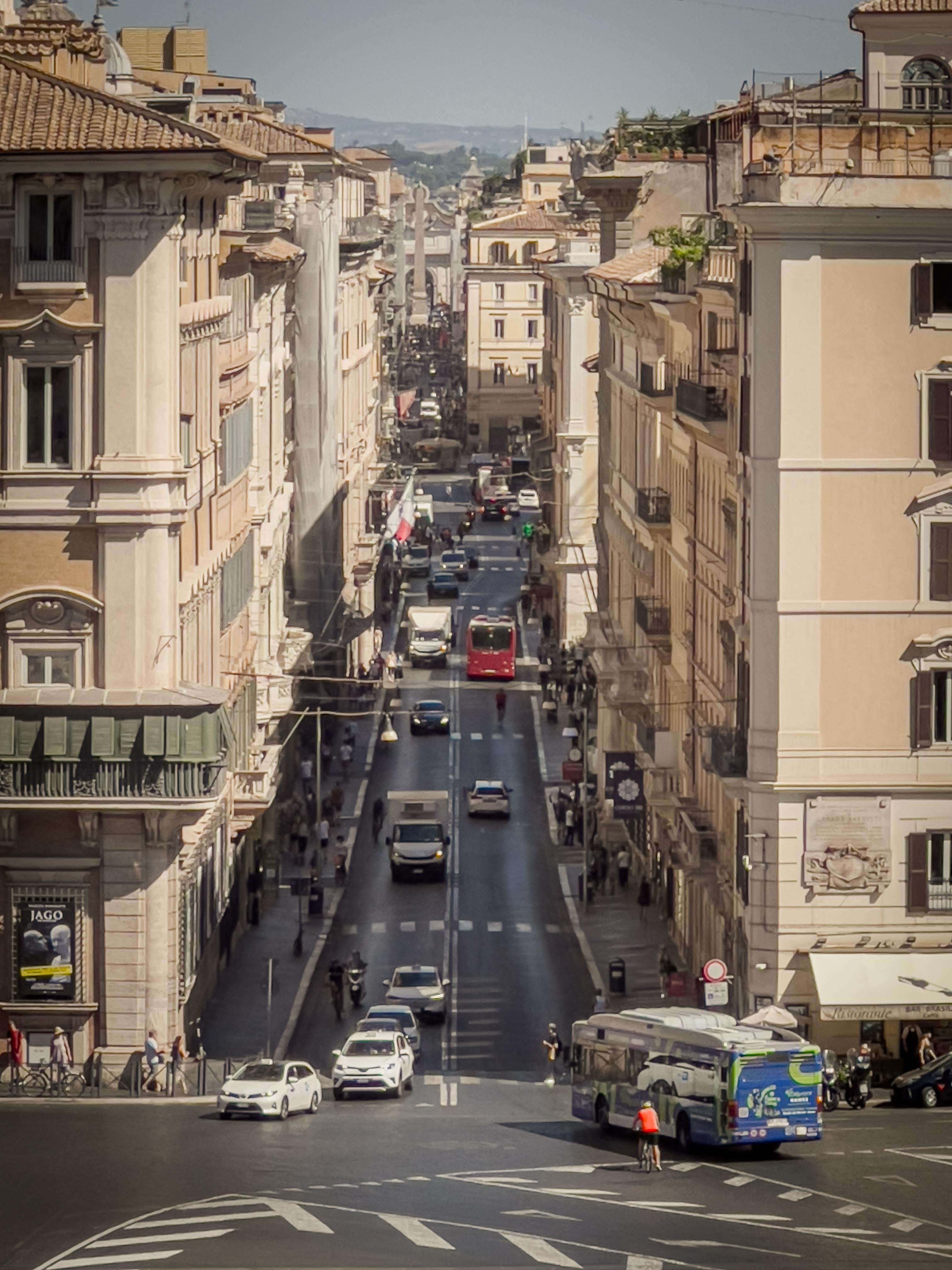 Strada trafficata in un contesto urbano, probabilmente in una città europea. La strada è fiancheggiata da edifici storici con dettagli architettonici classici. Ci sono veicoli tra cui auto, autobus e un ciclista. La prospettiva è dall’alto, offrendo una vista lungo tutta la lunghezza della strada. La scena è ben illuminata, suggerendo che potrebbe essere giorno.