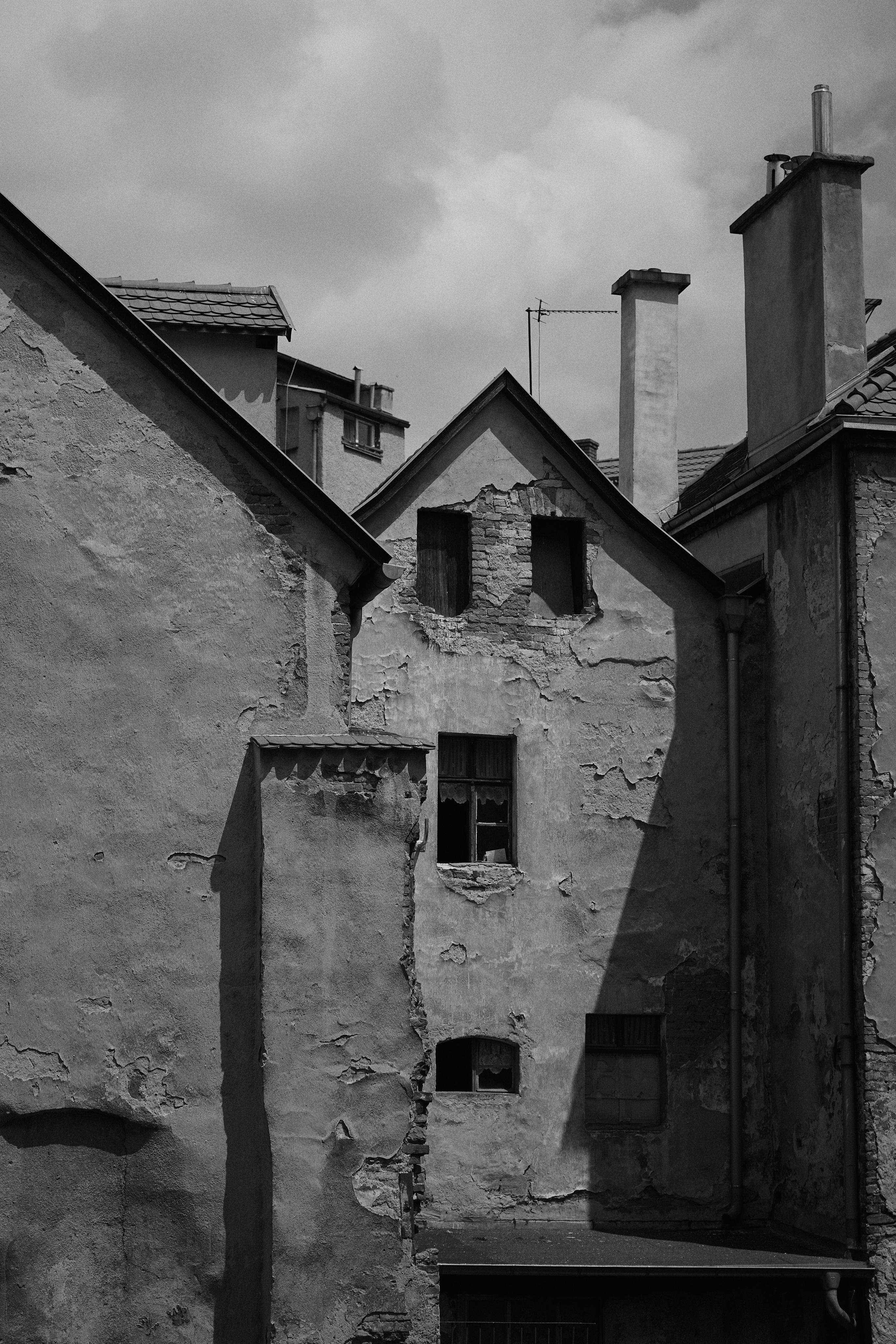A weathered old building with cracked and peeling plaster, featuring multiple small windows, some boarded up. The structure has a sloped, tiled roof and several large chimney stacks. Shadow and light contrast sharply against the façade, highlighting the building's textures. The sky in the background is overcast with partially cloudy conditions.