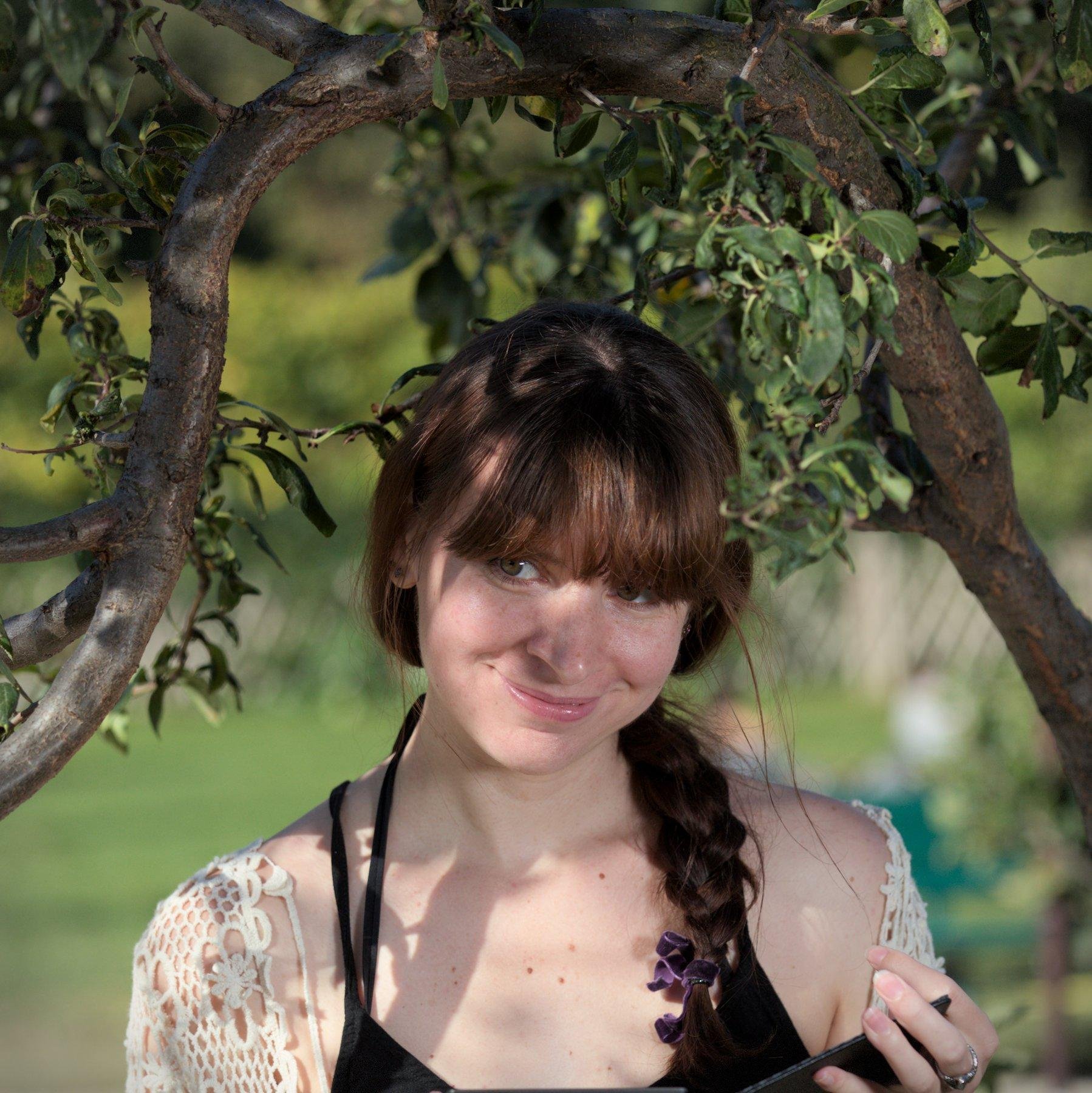 Photo carré. Gros plan d’une jeune femme châtain portant un débardeur noir et un gilet crocheté blanc aux motifs floraux, coiffée d’une natte qui pends par dessus son épaule gauche. Elle se trouve sous une branche d’arbre qui est tordu de manière à former un cadre au dessus de sa tête. Elle est souriante avec les yeux regardant en coin d’un air amusé