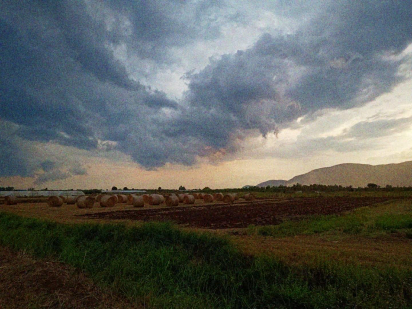 Paesaggio di campagna al tramonto, con balle di fieno, montagne in lontananza e cielo nuvoloso.
