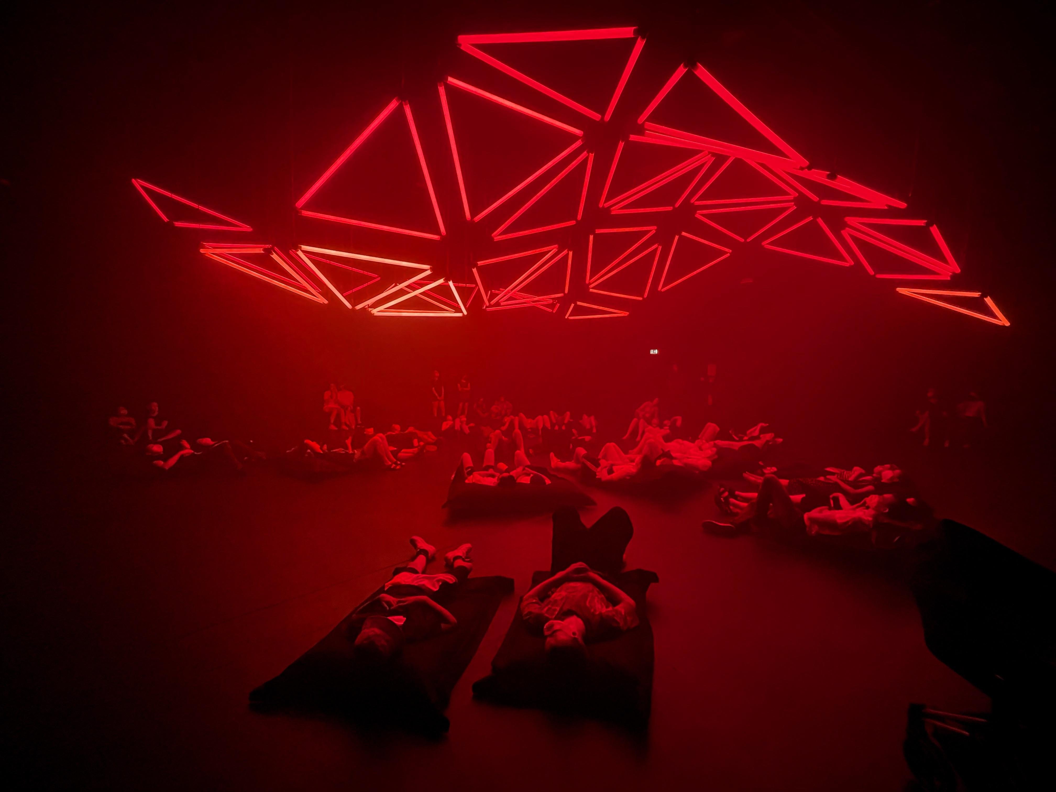 The image shows a room with people lying on the floor or on cushions, looking up at a ceiling light installation. The ceiling features an arrangement of red neon geometric shapes that form a visually striking pattern. The overall atmosphere appears immersive and artistic, as the part of an art installation or an event.