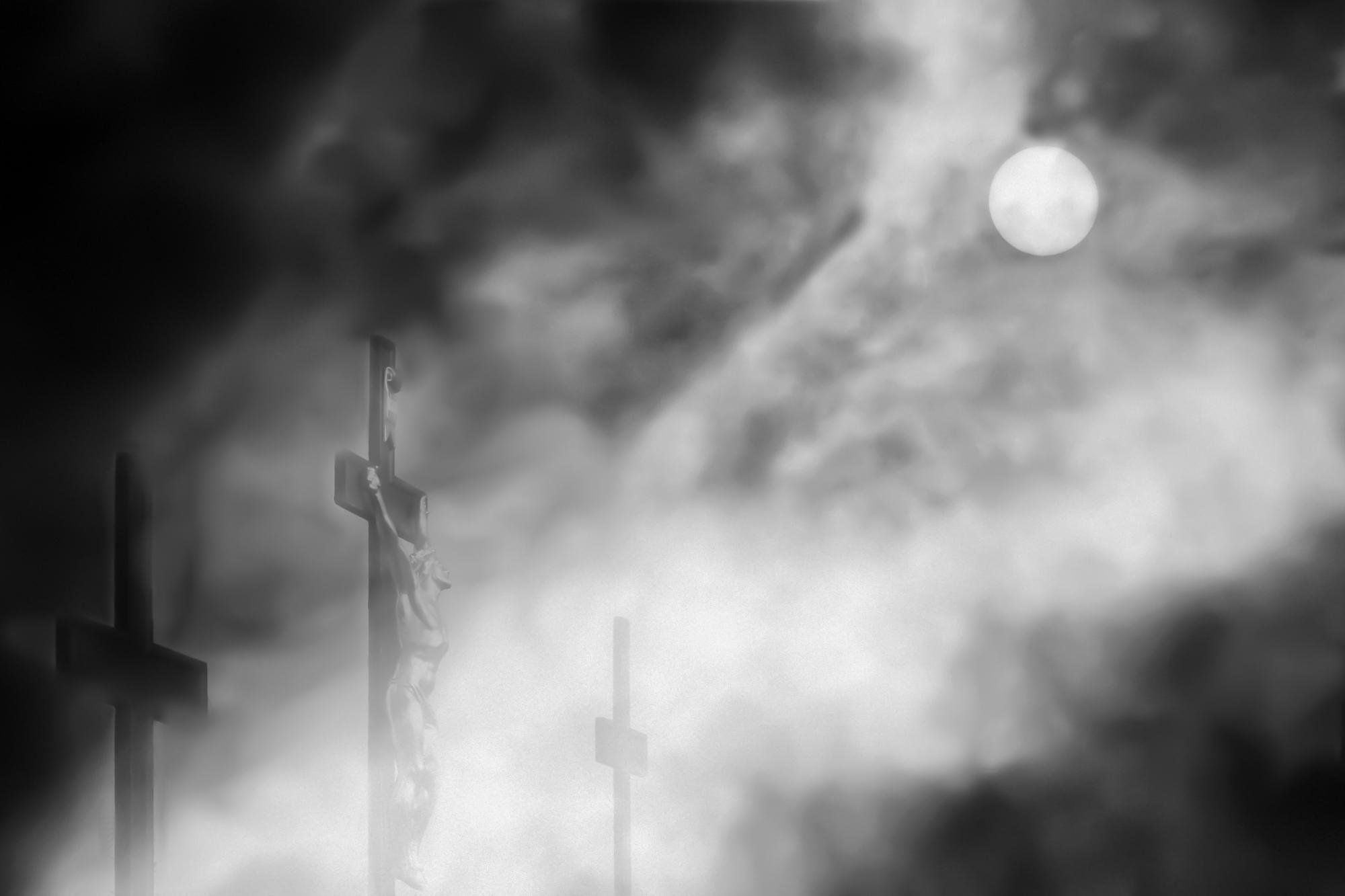 Three crosses emerging from dense fog, with the tallest cross in the left foreground and two smaller crosses in the background; a bright full moon visible in the upper-right amid swirling clouds in a black-and-white scene.