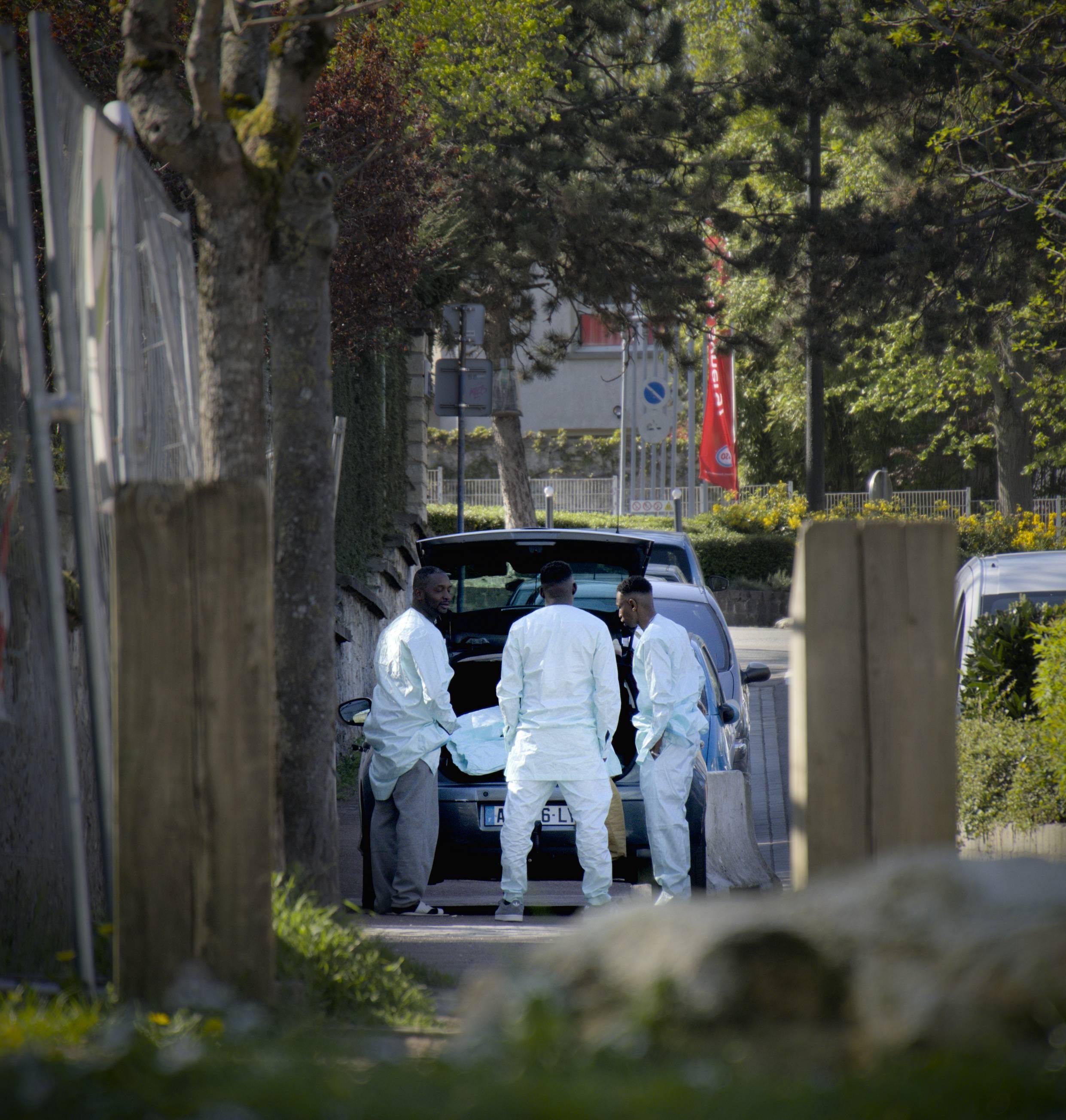 L'image montre trois personnes en combinaison blanche rassemblées autour d'une voiture dont le coffre est ouvert. Ils semblent engagés dans une activité ou une discussion.