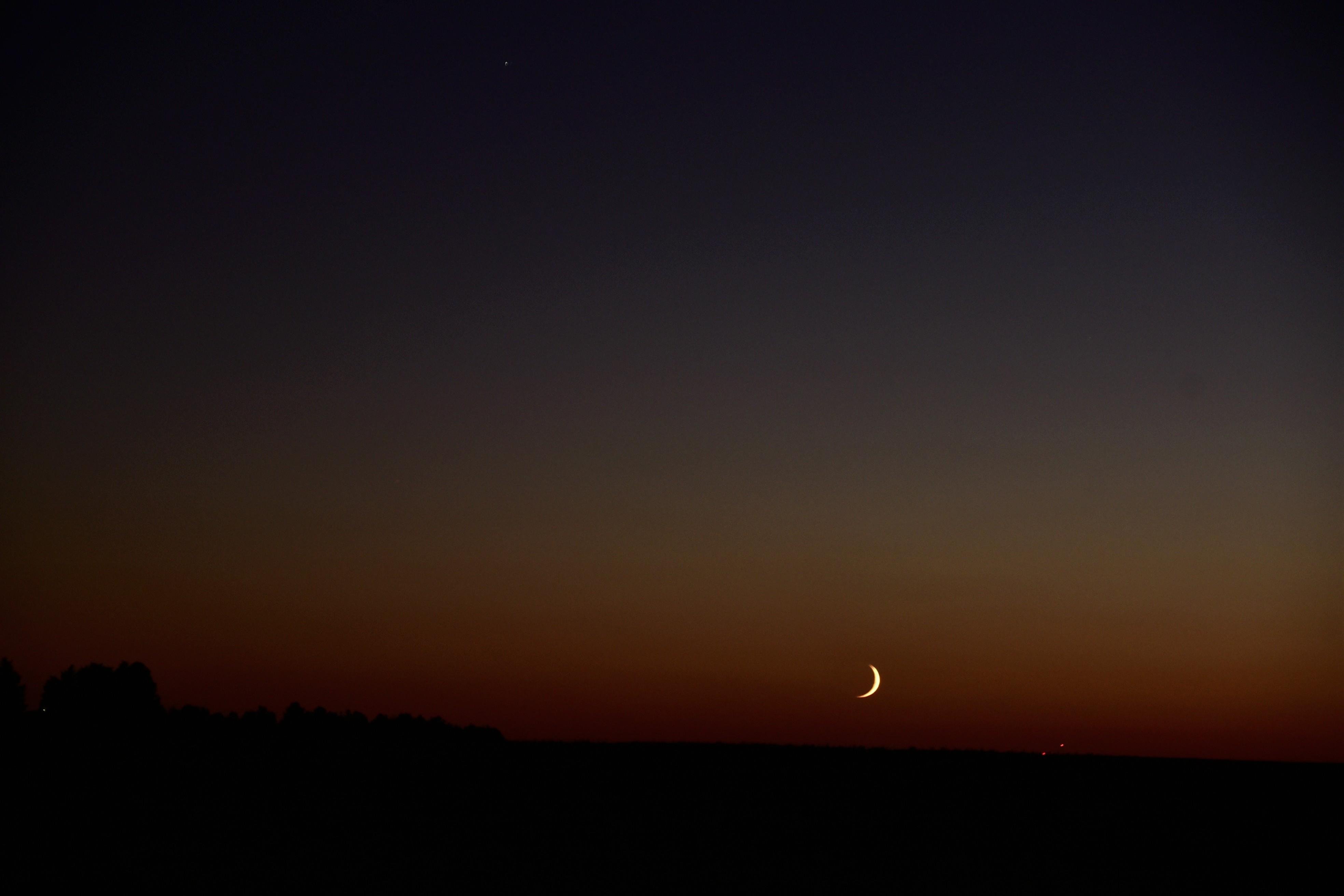 A thin orange crescent moon hangs low over the darkened plains against a deep blue sky that fades to reddish hues