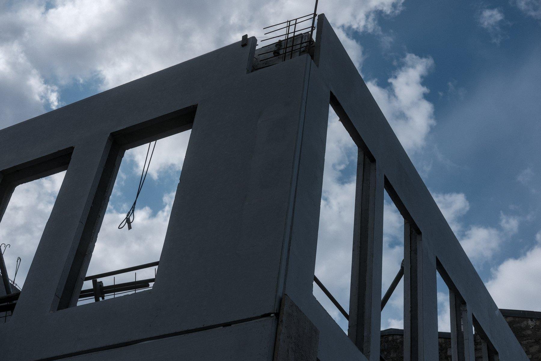Photo horizontale. Contre plongée sur un coin d’immeuble en construction. On voit des ouvertures pour de futures fenêtres, des fils qui pendent, un bout d’armature de béton, le ciel bleu nuageux à travers les ouvertures