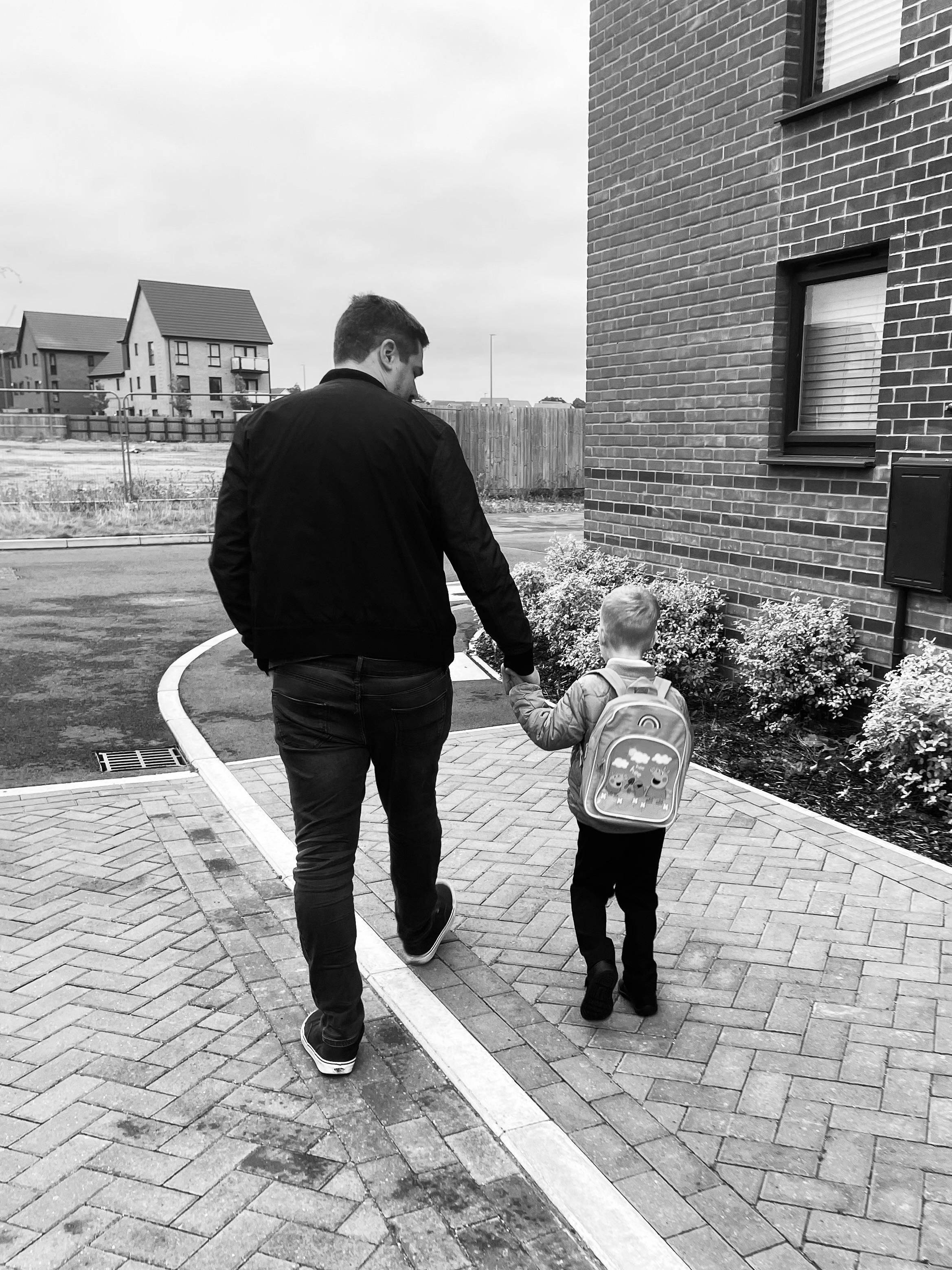 The image shows a black-and-white photo of an adult and a child walking along a paved path. The adult is holding the child's hand. The child is wearing a backpack. They are walking alongside a brick building, and there are residential houses in the background.
