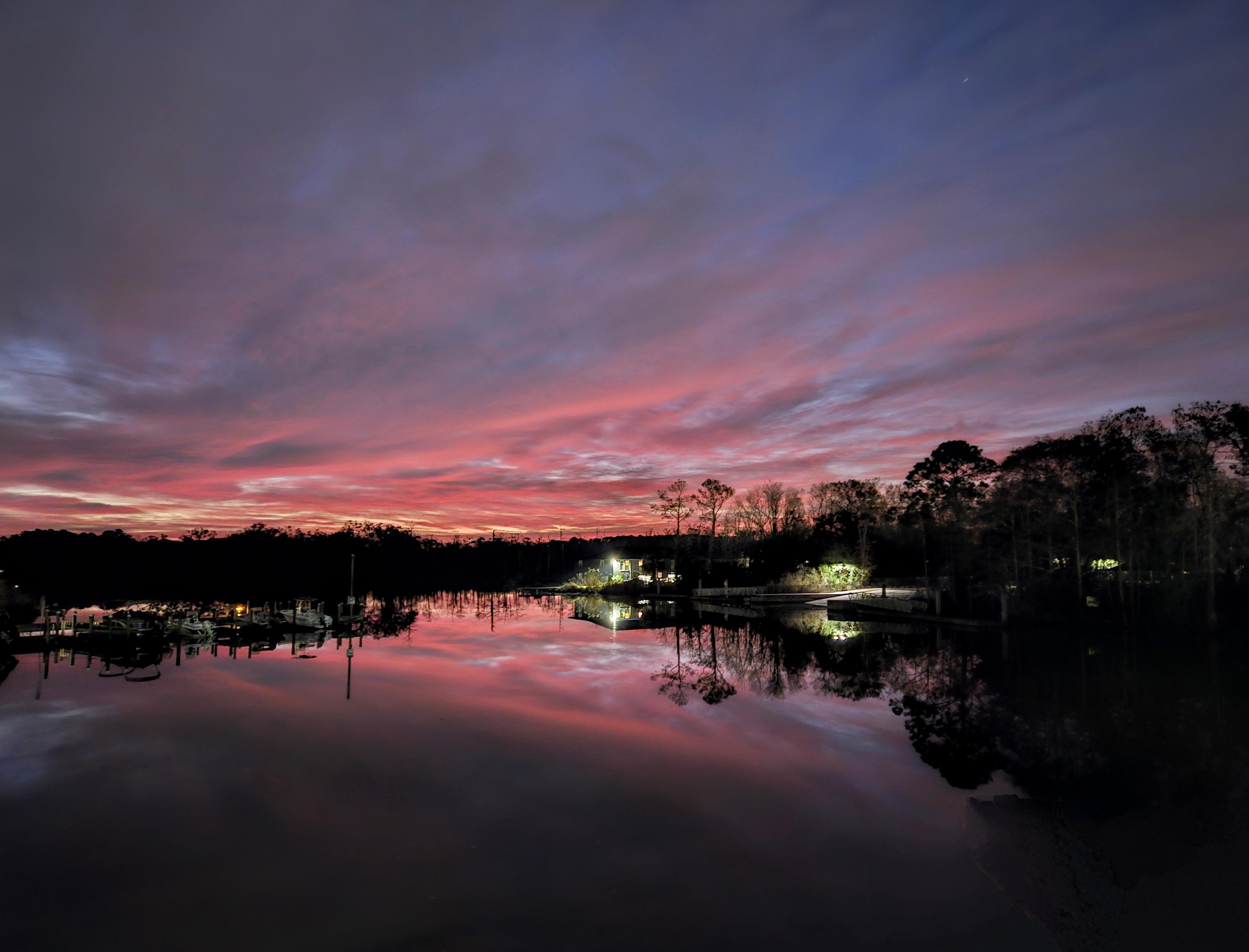 Sunset over a tranquil lake with vibrant pink and purple clouds reflecting on the water. Silhouetted trees line the horizon, and faint lights are visible near a dock on the right side, enhancing the serene atmosphere.