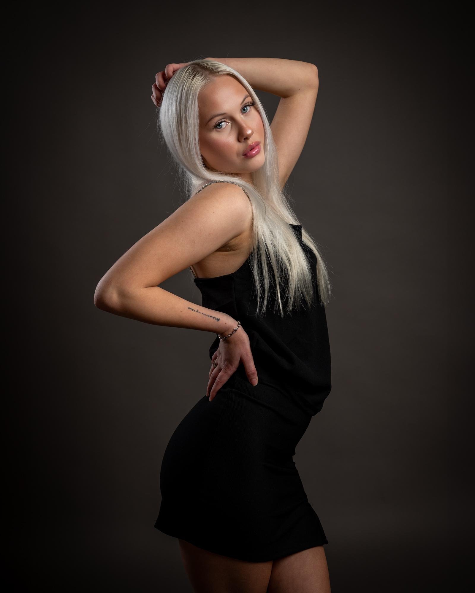 The image shows a person with long, light-colored hair posed against a plain, dark background. They are wearing a black sleeveless outfit and have one hand on their hip while the other is resting on their head. The lighting highlights their features, giving the photograph a professional studio look.
