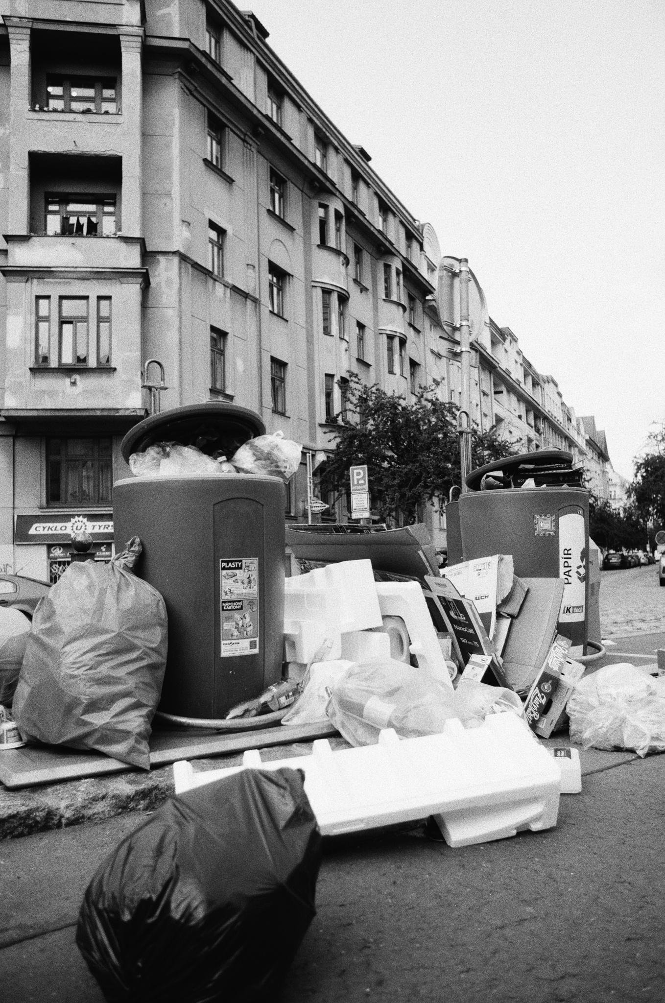 Overflowing street trash and recycling bins with plastic bags, cardboard, and foam packaging piled on the sidewalk, set against a row of multi-story apartment buildings along a city street.