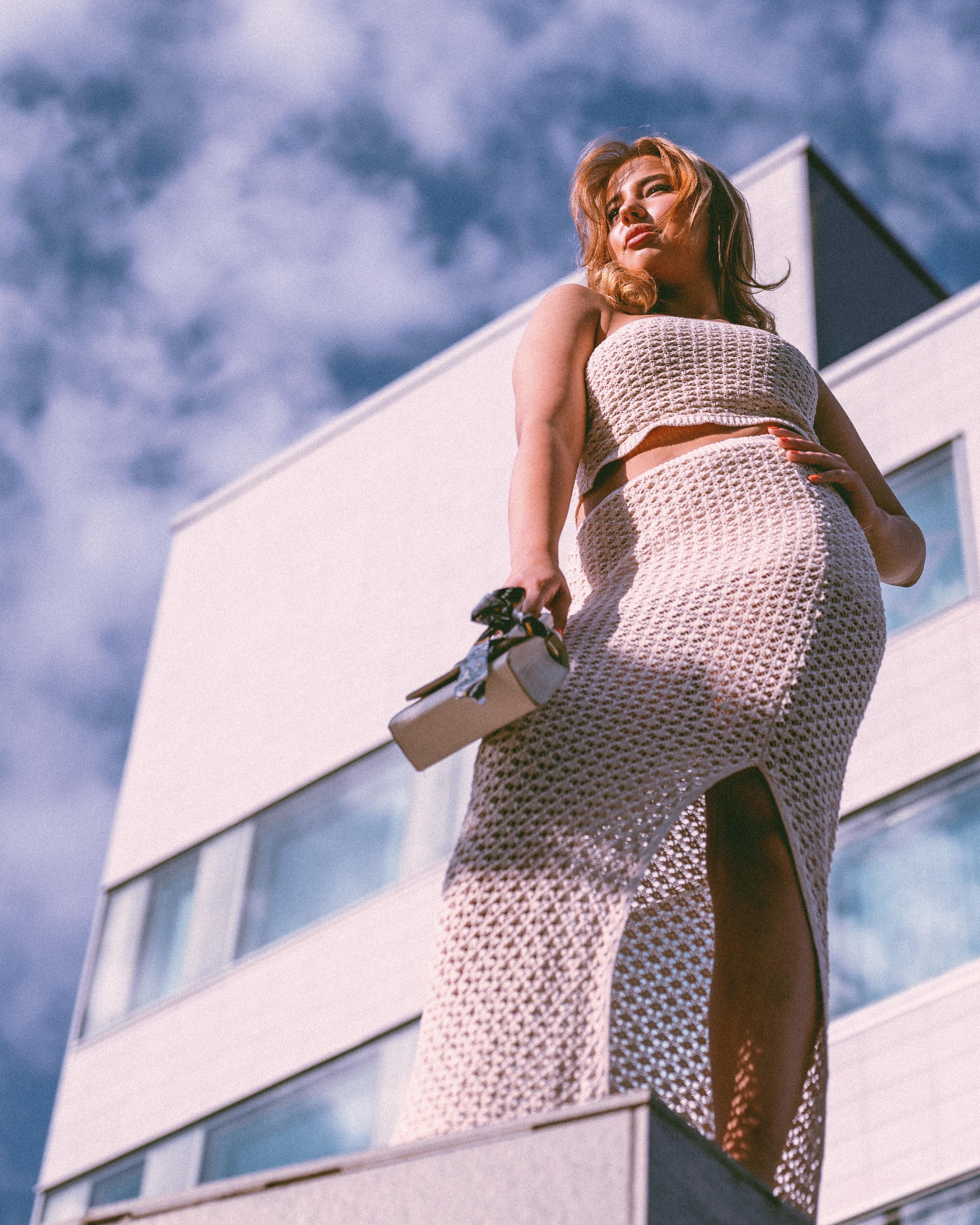 The image shows a person wearing a textured, light-colored two-piece outfit. They're standing on a ledge or platform in front of a modern building, looking upward. The sky is partly cloudy, and the perspective is from below, emphasizing the person against the backdrop of the building and sky.