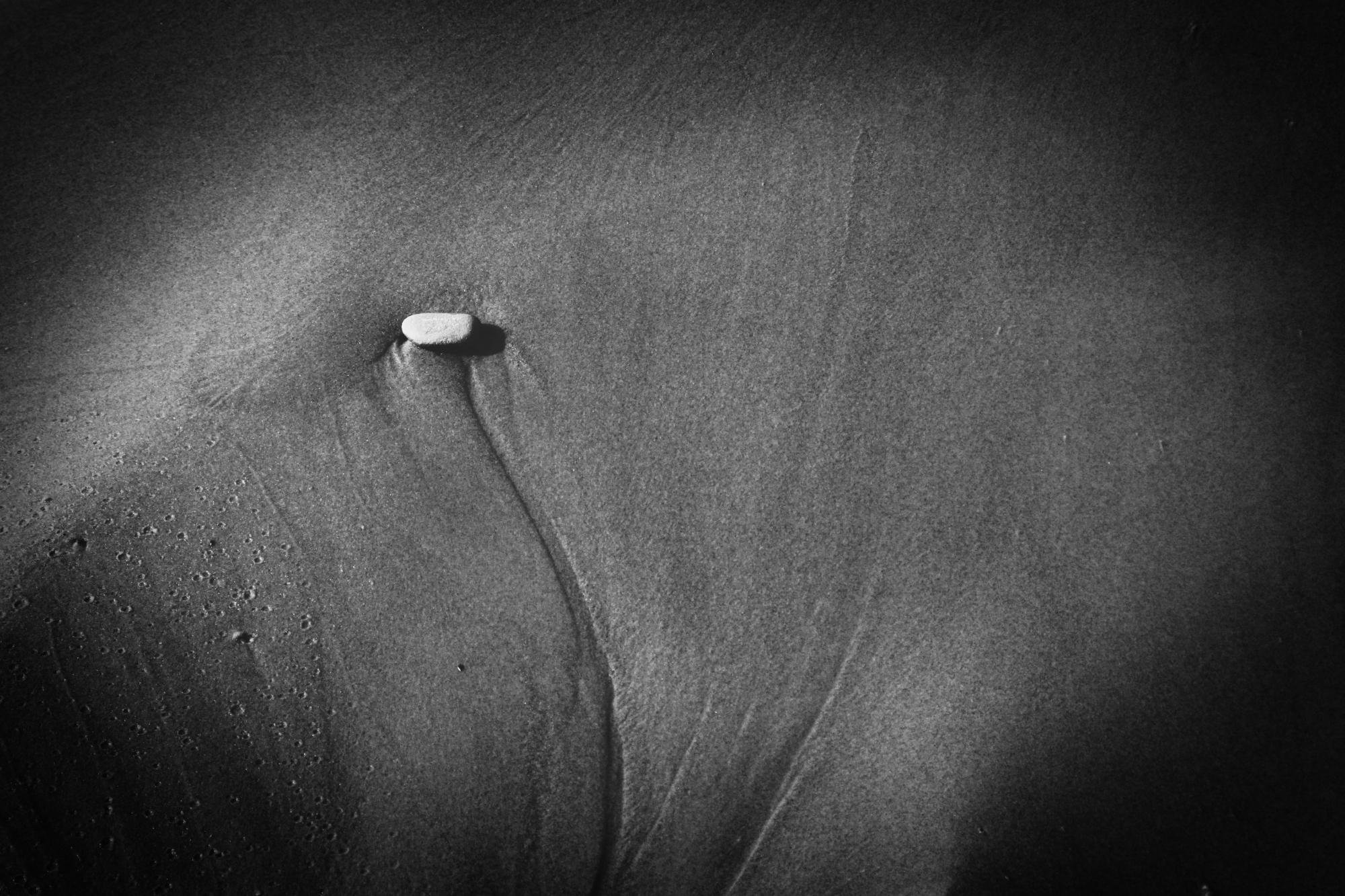 A black-and-white close-up of a sandy beach, with a single pebble lying in the upper left corner, from which a thin groove runs downward across the surface.