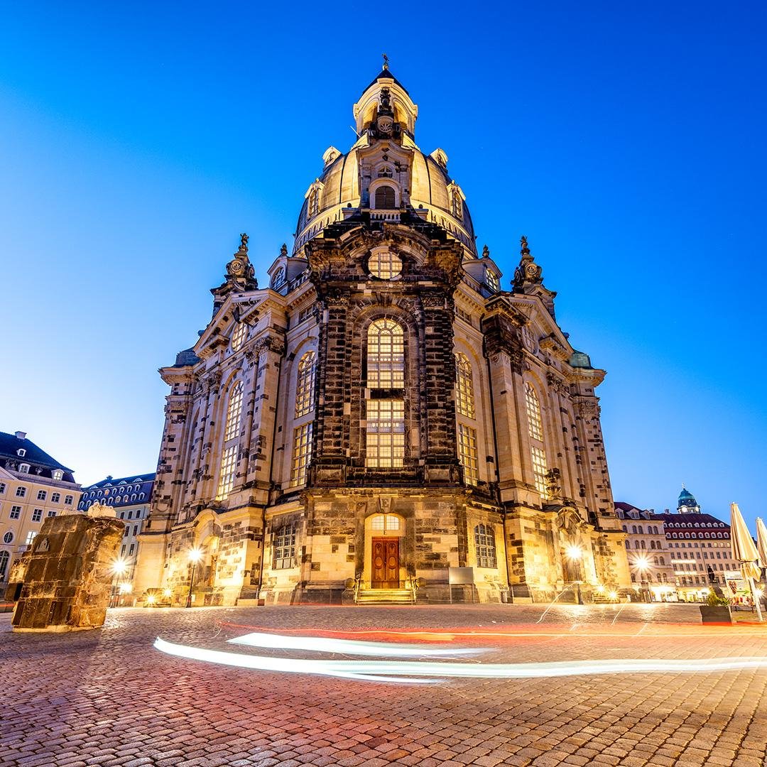 Die beleuchtete Frauenkirche in Dresden zur blauen Stunde, fotografiert frontal aus niedriger Perspektive. Das barocke Gebäude mit großer Kuppel und hohen Fenstern steht auf einem Kopfsteinpflasterplatz, während im Vordergrund durch eine Langzeitbelichtung Lichtspuren vorbeifahrender Fahrzeuge zu sehen sind. Der Himmel ist tiefblau und die warme Beleuchtung hebt die Architektur der Kirche hervor.